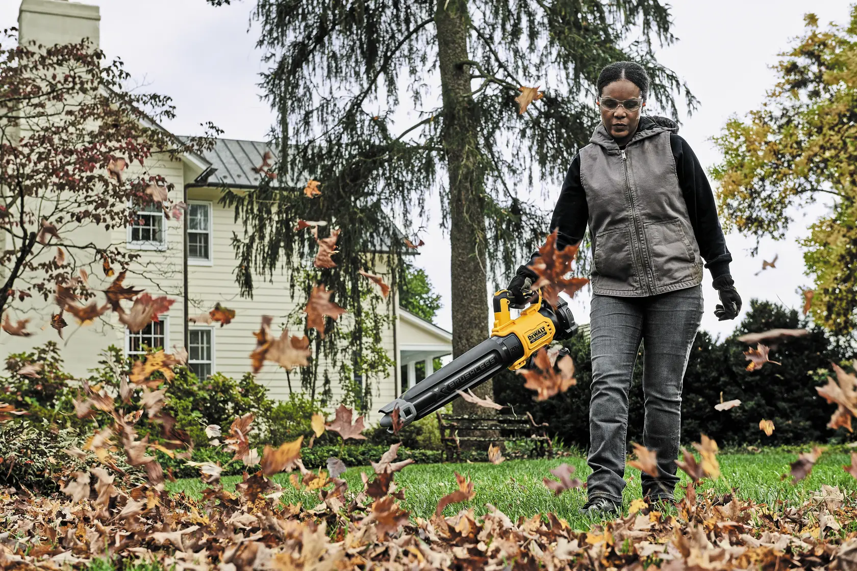 A person is using a yellow DEWALT leaf blower to clear fallen leaves on a lawn in front of a house. The person's face is blurred for privacy.