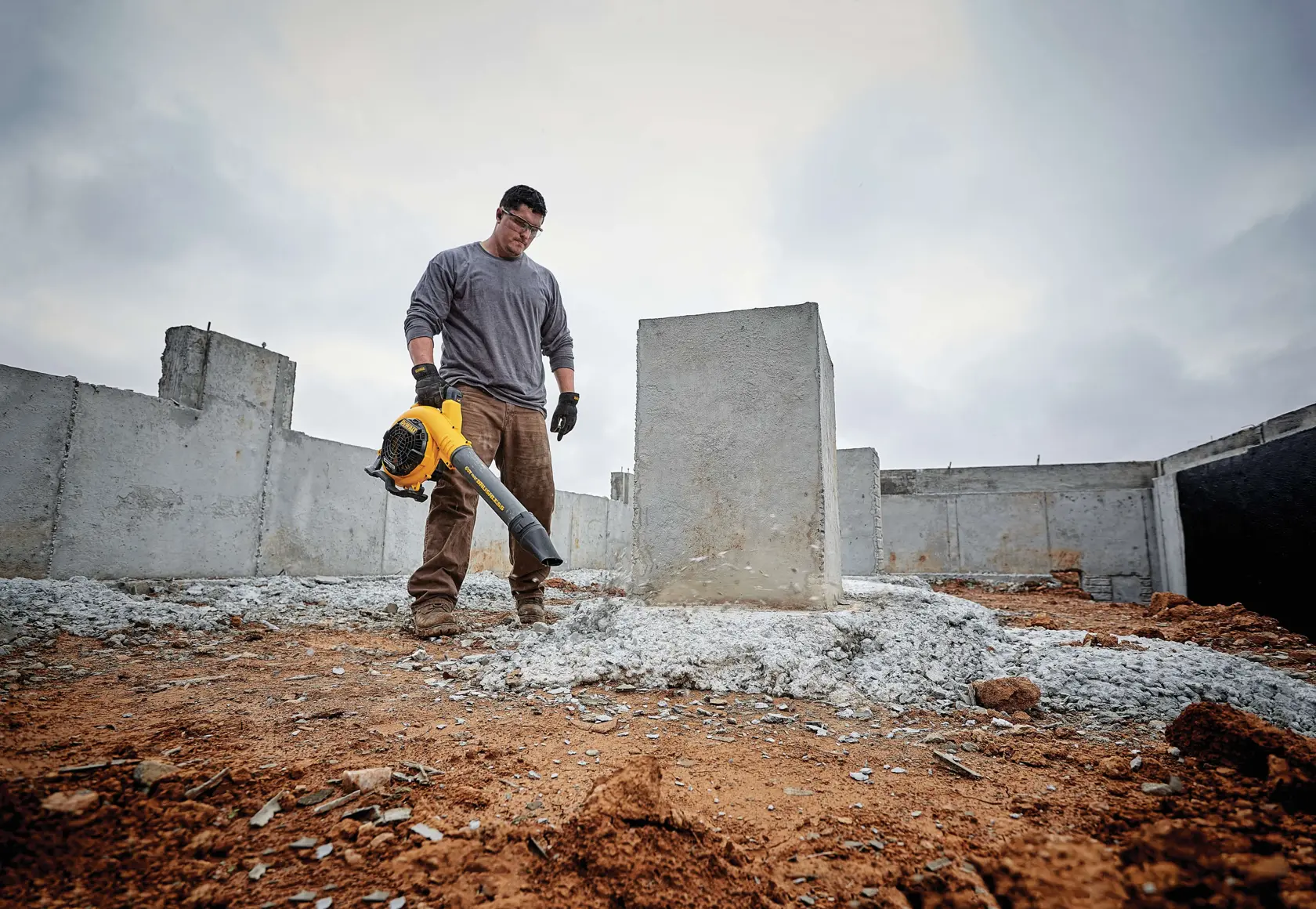 A person wearing work clothes and gloves uses a yellow DEWALT blower to clear debris around concrete blocks at a construction site. The sky is cloudy and the ground is covered with rubble and dirt.