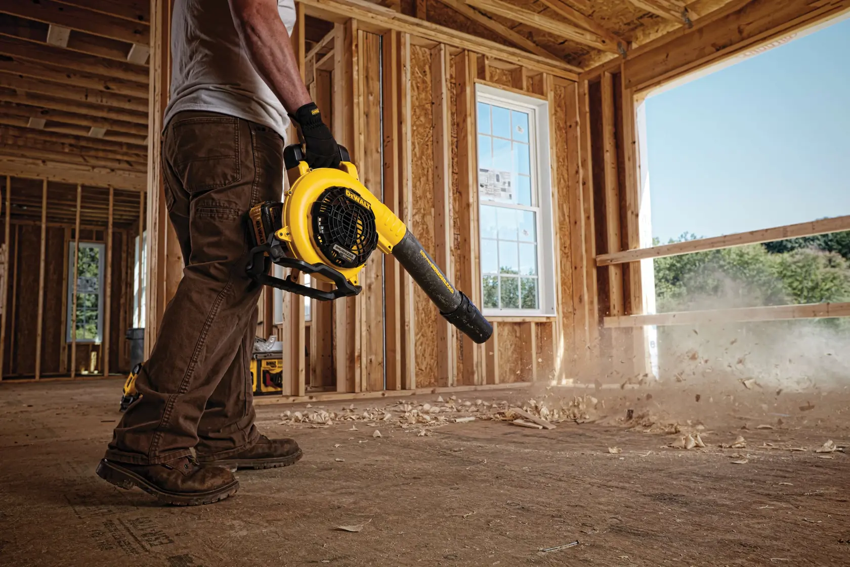 A person is using a yellow DEWALT cordless blower to clear debris from the floor of a wooden construction site.