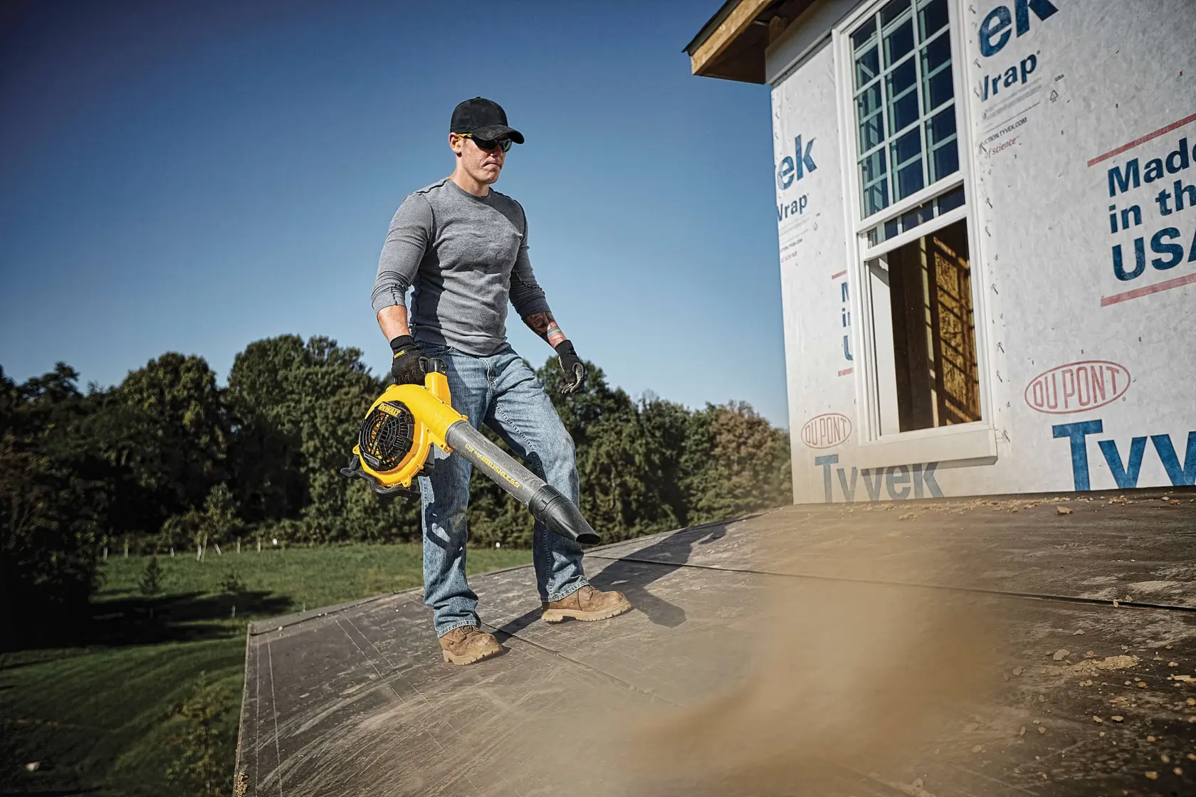 A person wearing gloves and a cap is using a yellow DEWALT leaf blower to clear debris from a roof at a construction site. The building is covered in Tyvek wrap, and there are trees and grass in the background.
