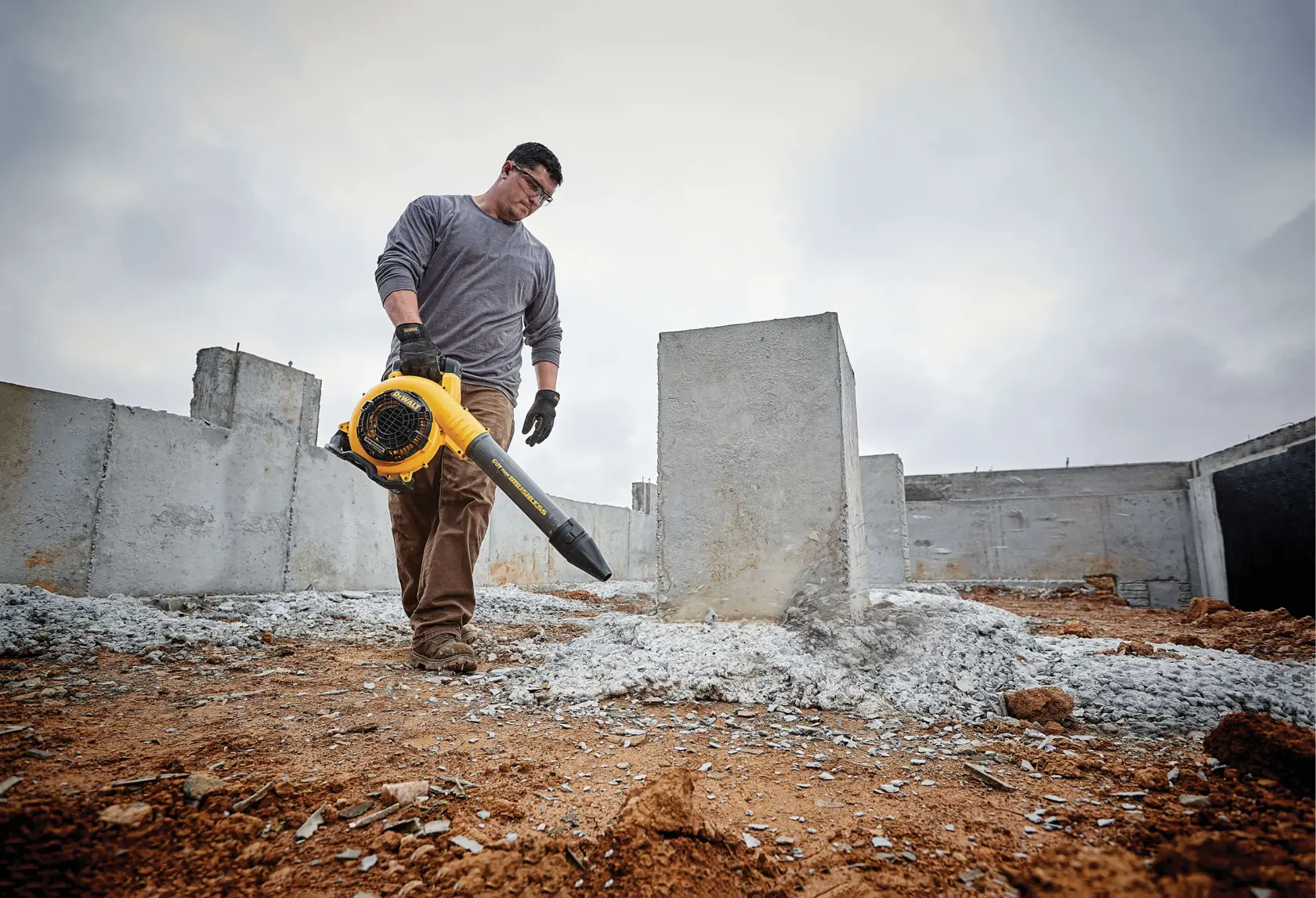 A person wearing work gloves uses a yellow DEWALT cordless blower to clear debris from a construction site with concrete structures and dirt ground.