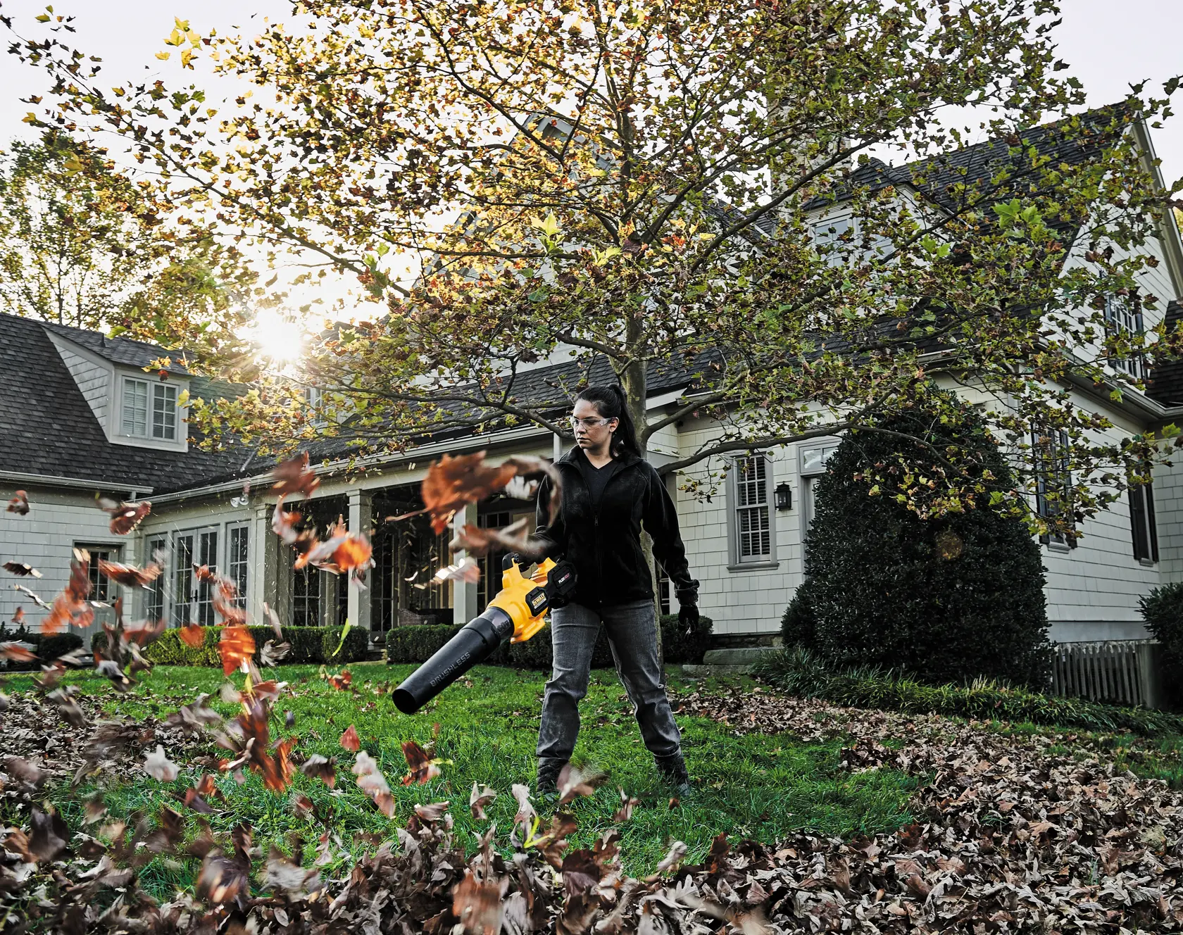 A person using a DEWALT leaf blower outdoors to move fallen leaves on a grassy lawn, with a house and trees in the background.
