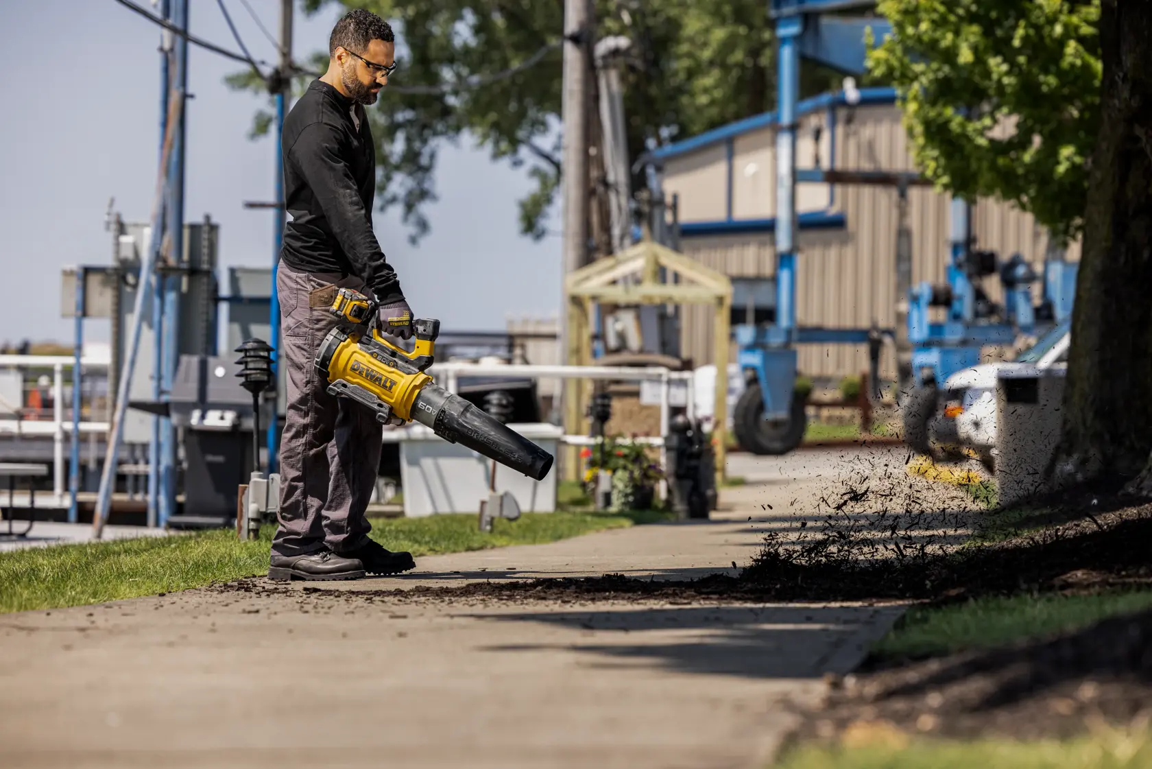 A person is using a yellow DEWALT cordless leaf blower to clear debris from a sidewalk outdoors. Industrial buildings and green trees are visible in the background.