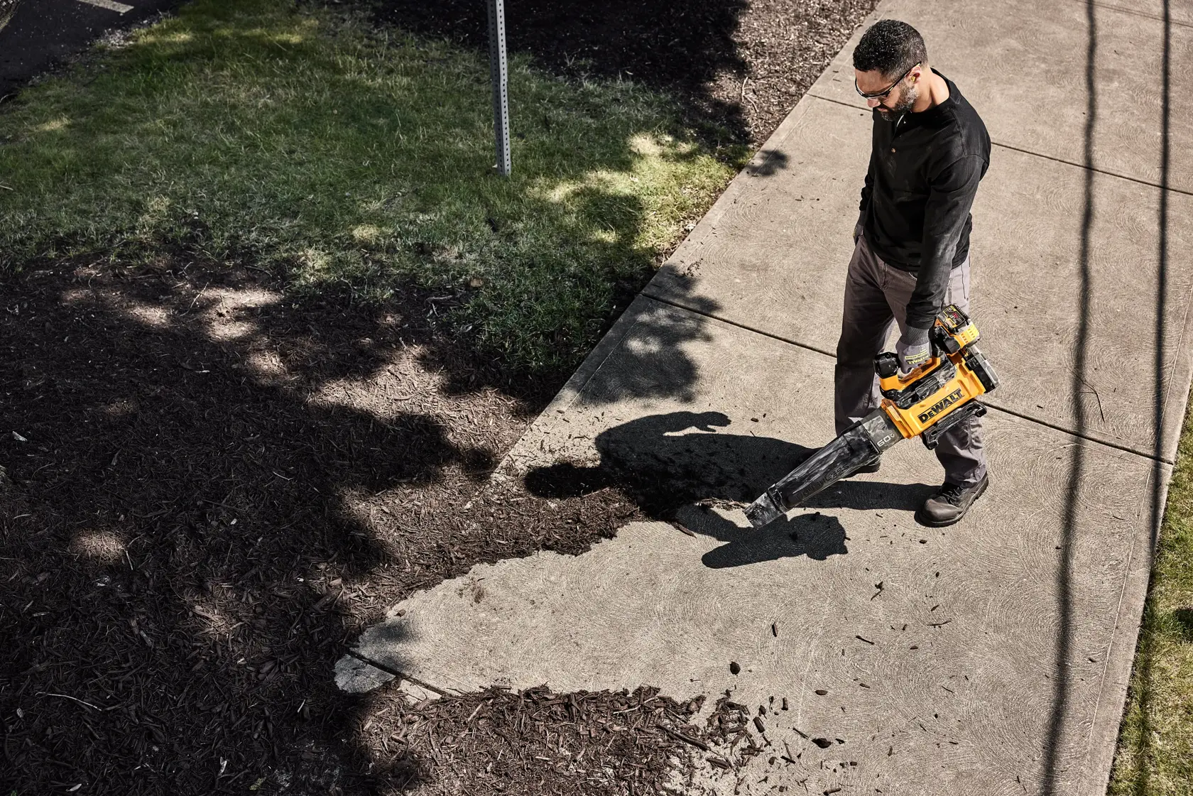 A person using a yellow and black DEWALT leaf blower to clear debris from a concrete sidewalk near a grass and mulch area.