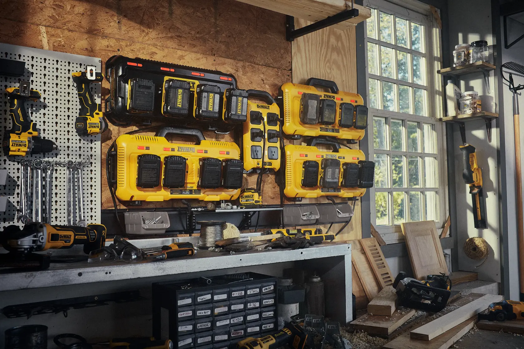 A workshop with several DEWALT battery chargers mounted on the wall, various DEWALT cordless power tools, and accessories organized on a workbench and pegboard. The room has large windows letting in natural light.