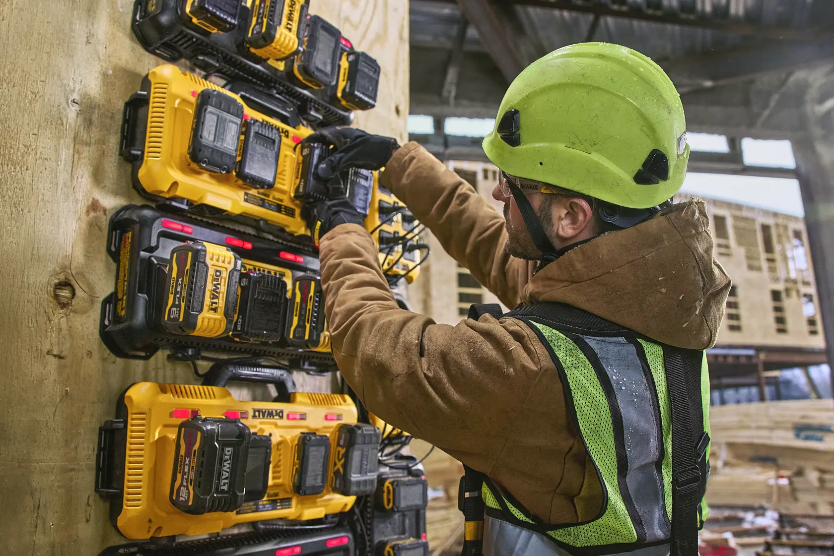A person wearing a green hard hat, gloves, and a safety vest is working with a wall-mounted DEWALT battery charging station at a construction site. Multiple yellow and black DEWALT chargers and batteries are attached to the wooden wall.