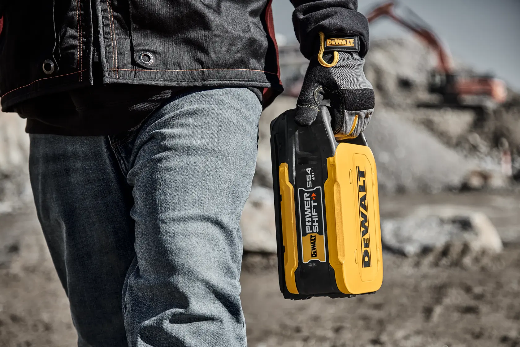 A person wearing work gloves and jeans holds a yellow and black DEWALT Power Shift 554 battery pack at a construction site with heavy machinery in the background.
