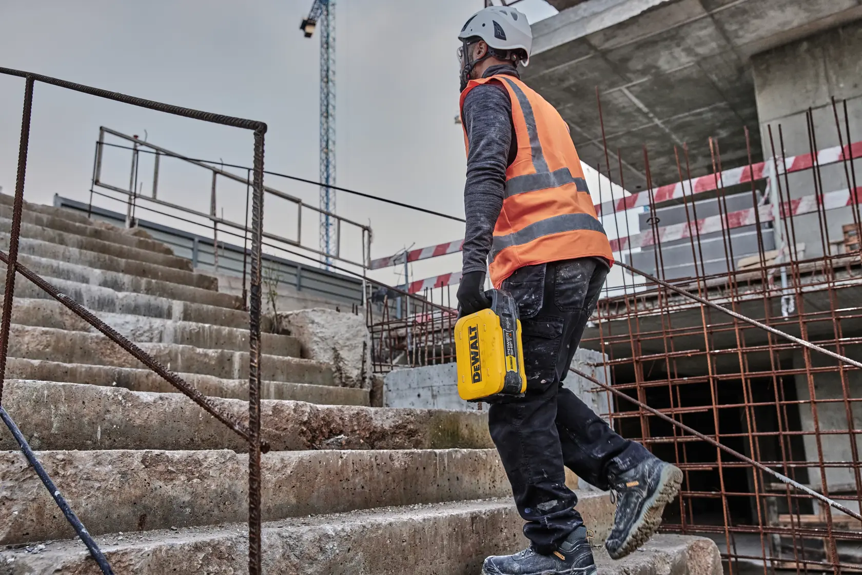 A person wearing safety equipment and an orange vest is walking up outdoor concrete stairs at a construction site, holding a yellow DEWALT tool.