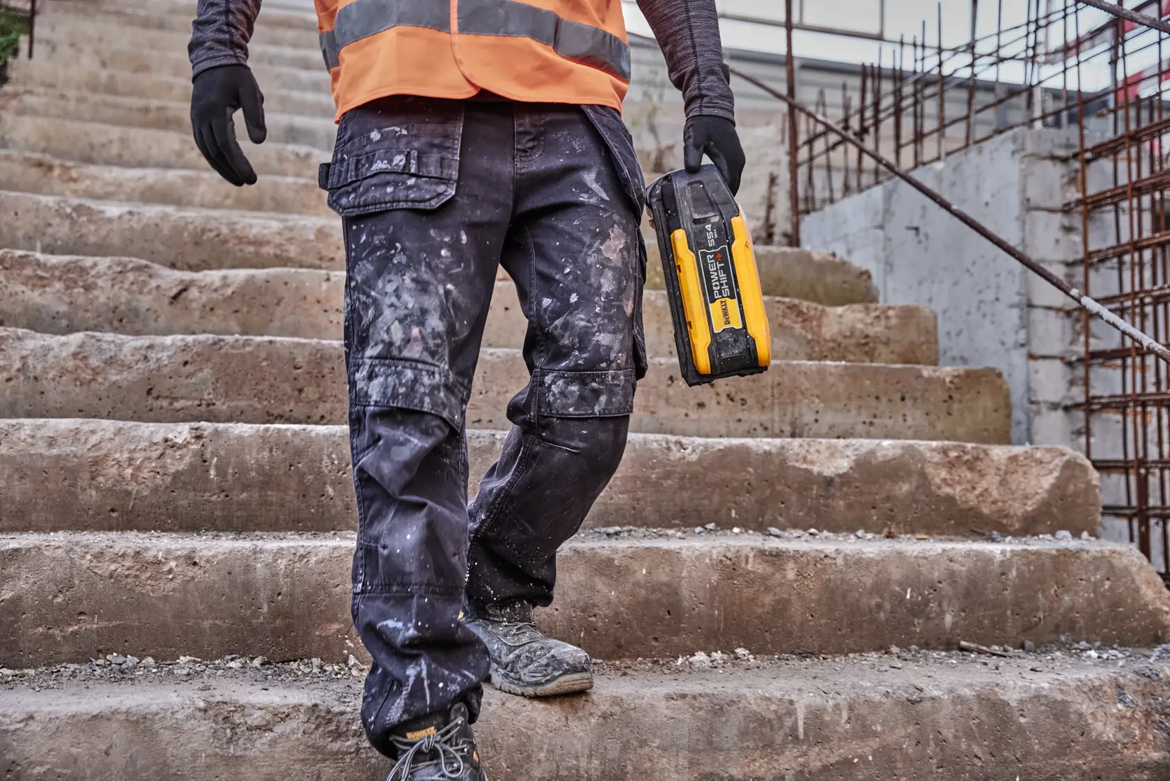 A person wearing work gloves and an orange safety vest walks down concrete steps at a construction site, holding a yellow and black DEWALT tool.