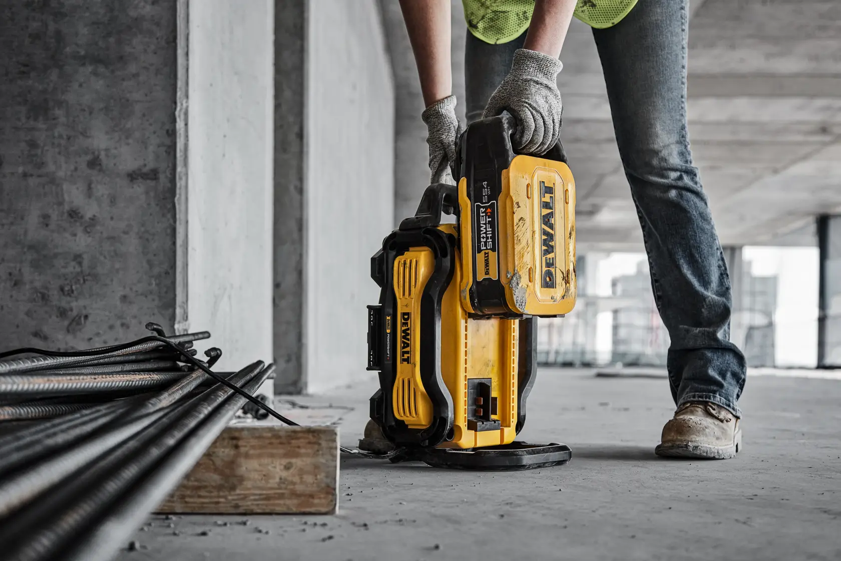 A person wearing gloves and work boots is placing a yellow DEWALT portable power station on a concrete floor at a construction site, near stacks of metal rods.