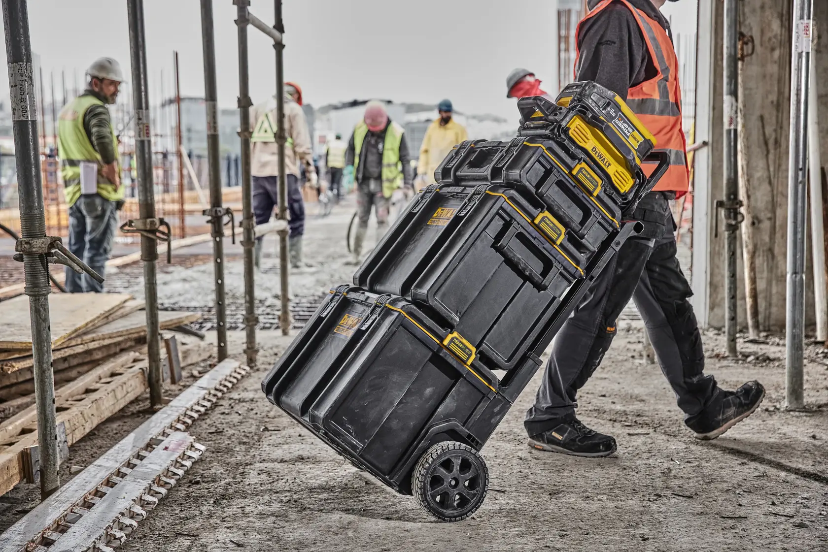 A person wearing a high-visibility vest is pulling a stack of black DEWALT toolboxes with yellow handles on a wheeled trolley at a construction site. Several workers and construction materials are visible in the background.