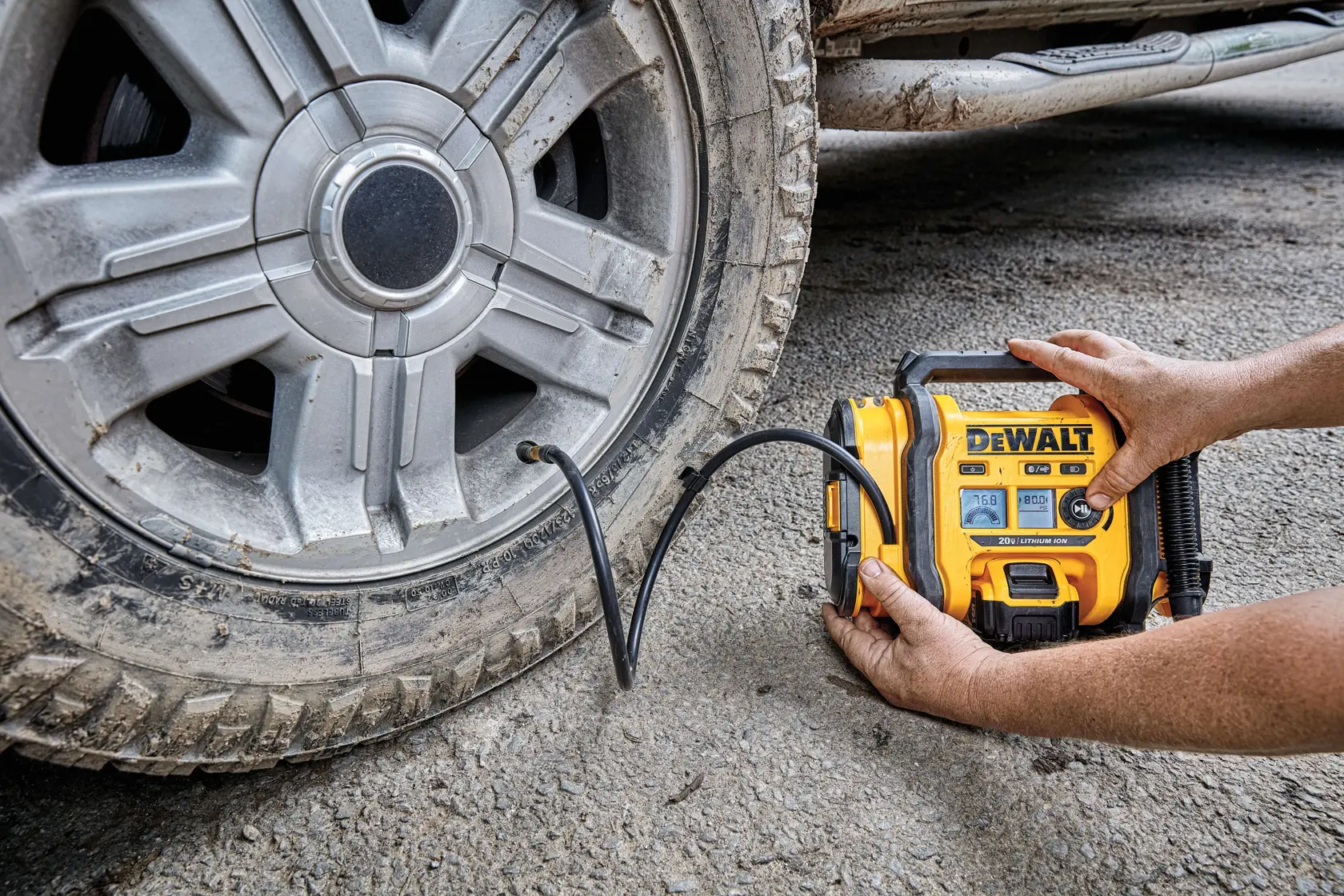 A person is using a yellow DEWALT portable air compressor to inflate a car tire on a rough, outdoor surface. The compressor’s digital display and hose are visible.