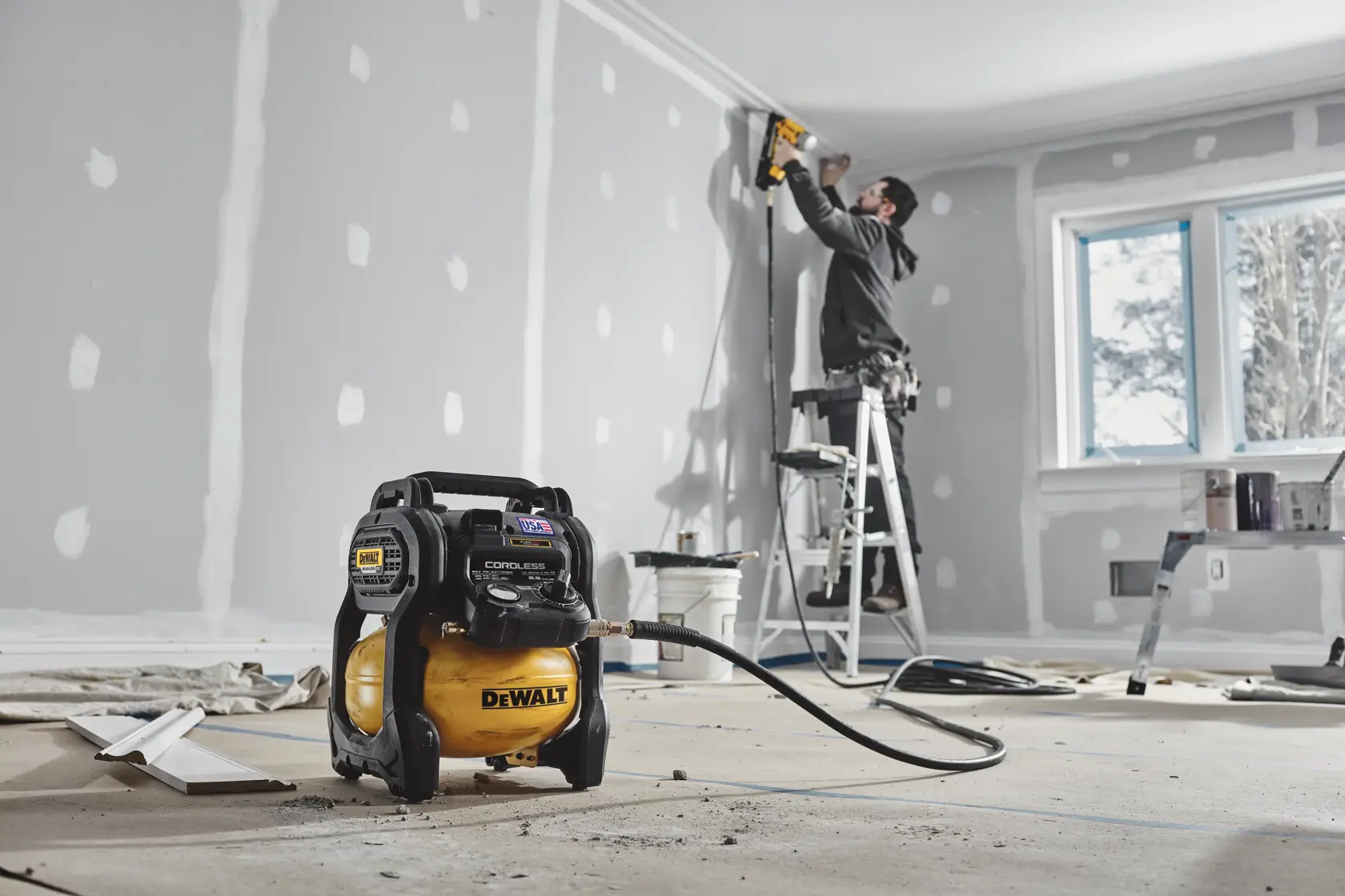 A DEWALT cordless air compressor is placed on the floor in the foreground of a room under construction. In the background, a person stands on a ladder using a power tool to work on the ceiling near the wall, which is covered in drywall with visible joint compound patches.