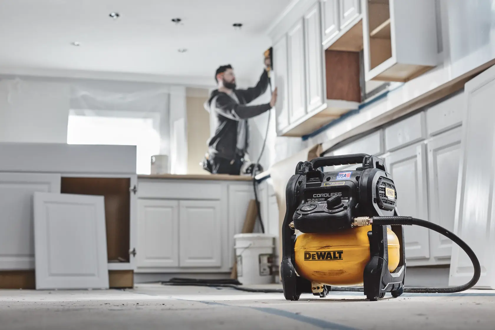 A DEWALT cordless air compressor (model DCC2520T1) is on the floor in a kitchen under renovation. Cabinets are being installed and a person blurred in the background is working with a power tool.