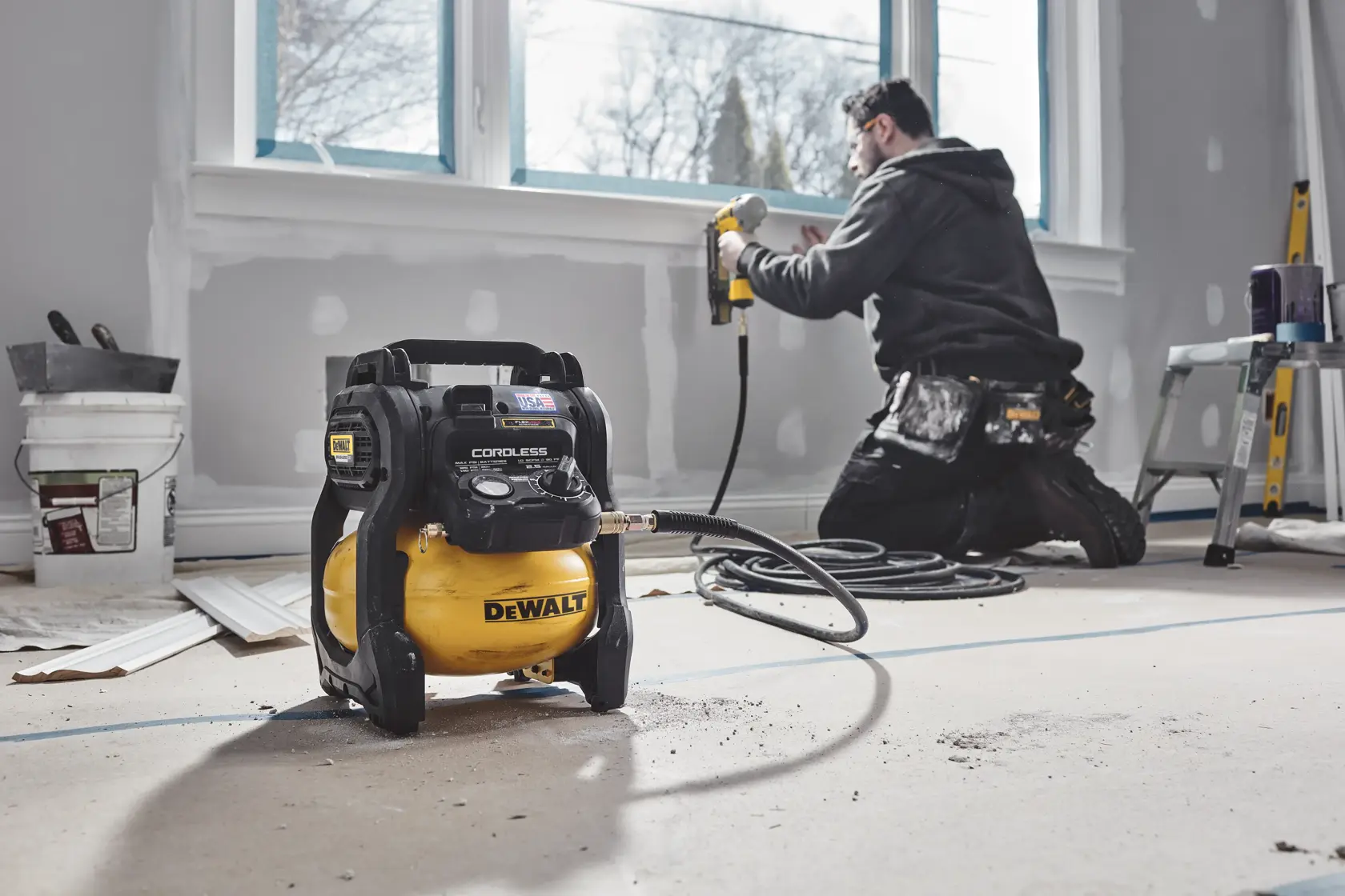 A DEWALT cordless air compressor is placed on the floor in a room under renovation. In the background, a person is kneeling and using a power tool near a window, surrounded by renovation materials and equipment.