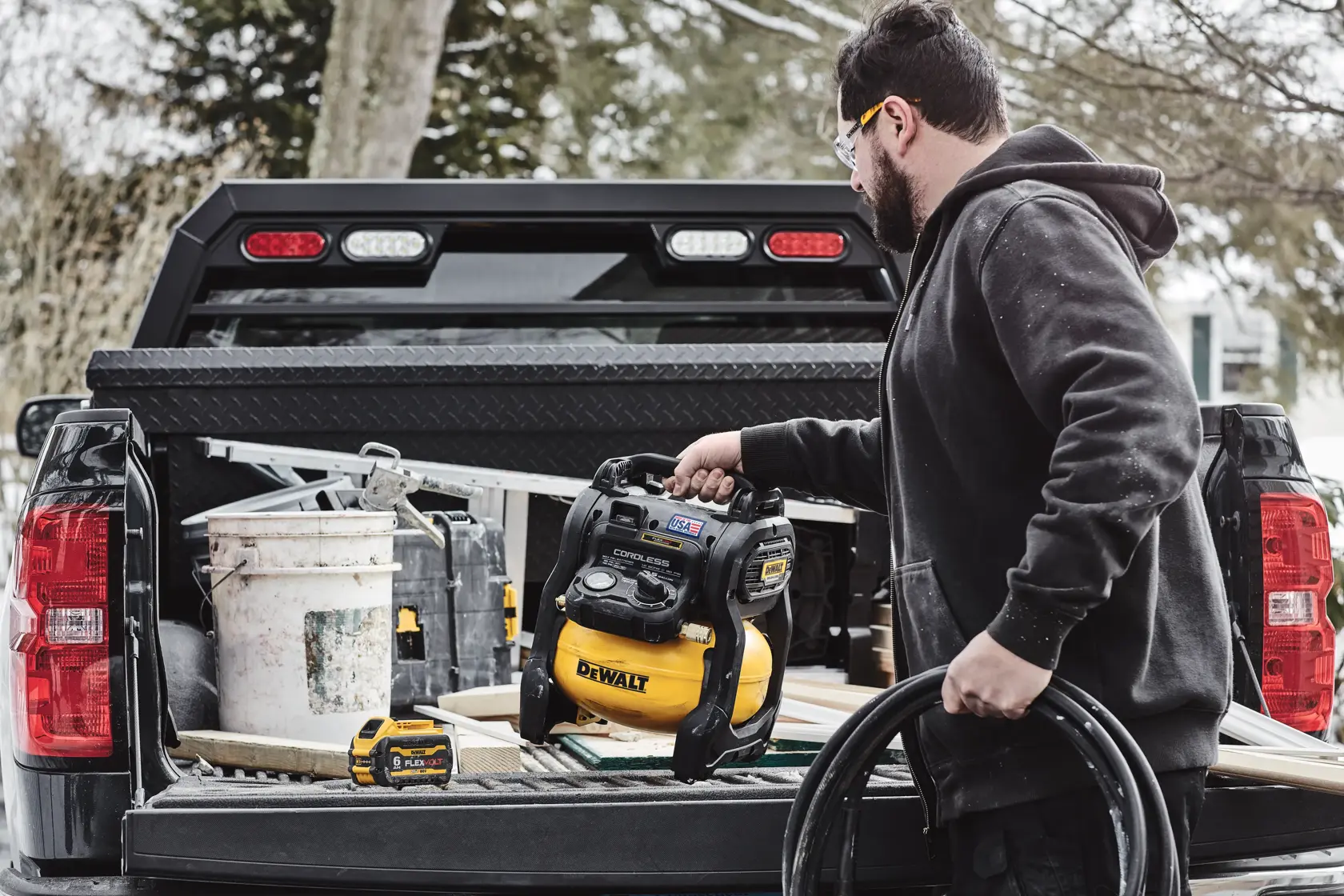 A person with a blurred face places a DEWALT cordless air compressor in the bed of a pickup truck, surrounded by construction tools and materials, including a large bucket, a hose, and a battery.