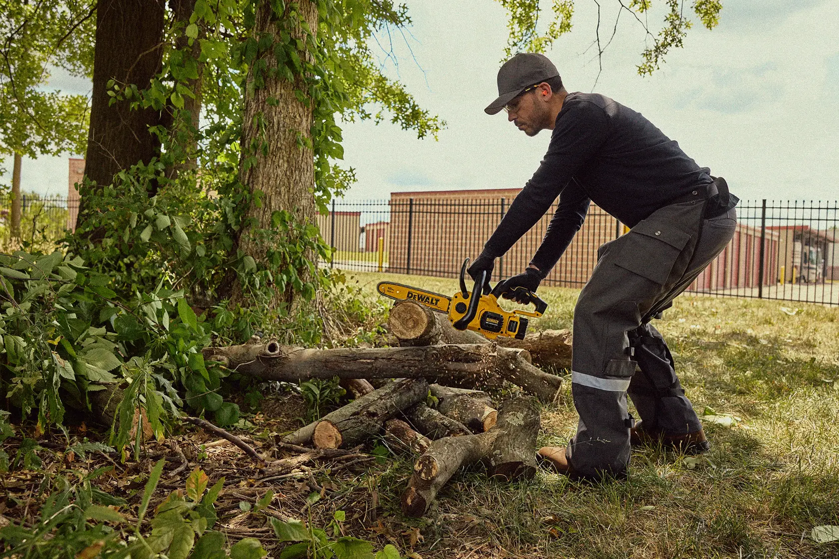 A person using a DEWALT cordless chainsaw to cut logs outdoors. The individual is wearing protective clothing and gloves, and appears to be working in a grassy area near trees and some buildings.