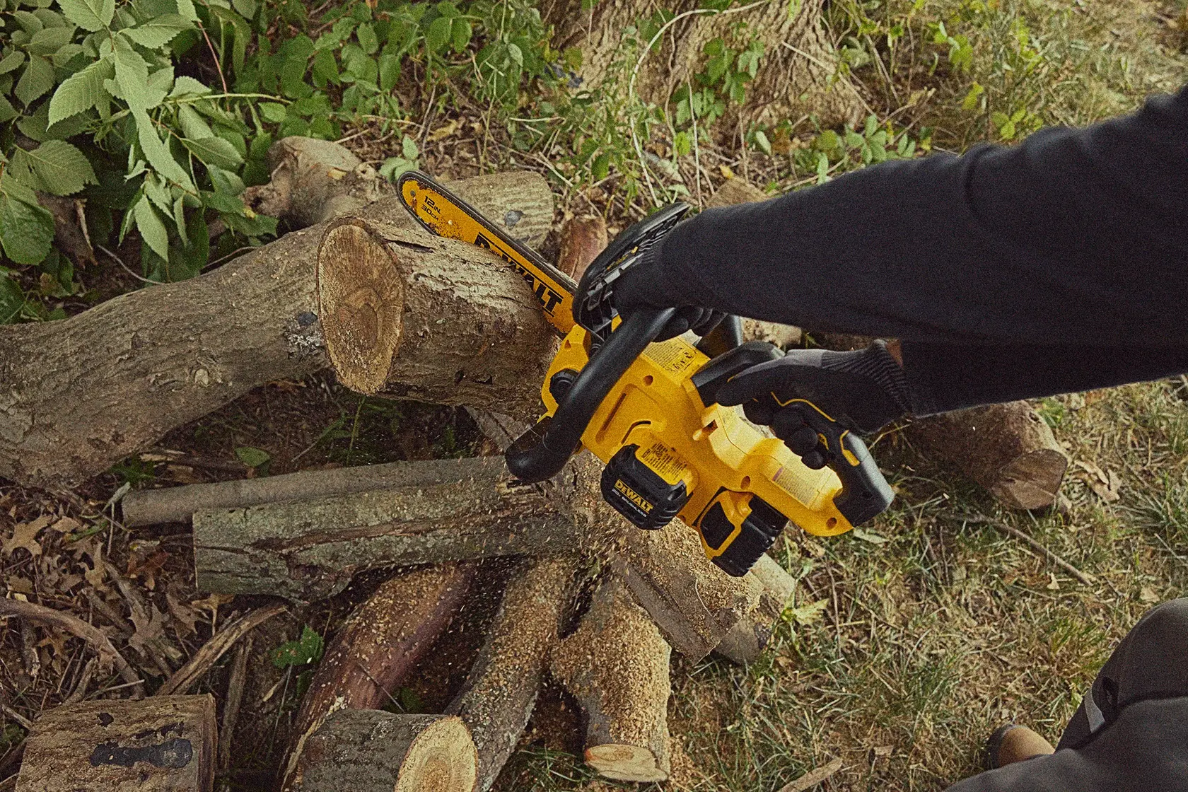 A person wearing black gloves uses a yellow DEWALT cordless chainsaw to cut a log outdoors. Several logs and green foliage are visible on the ground.