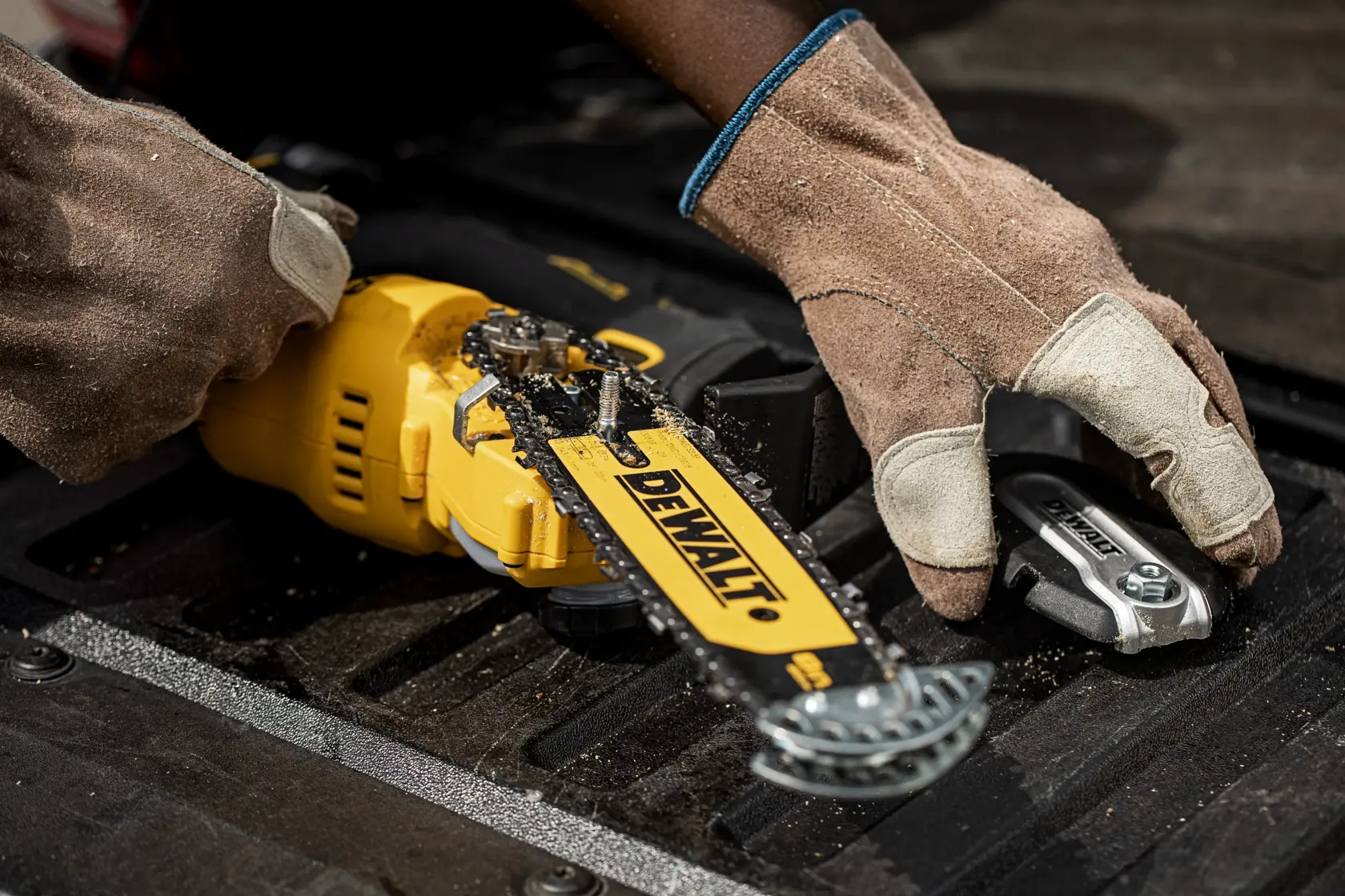 Close-up of a person wearing work gloves handling a yellow DEWALT cordless chainsaw and tools on a black surface.