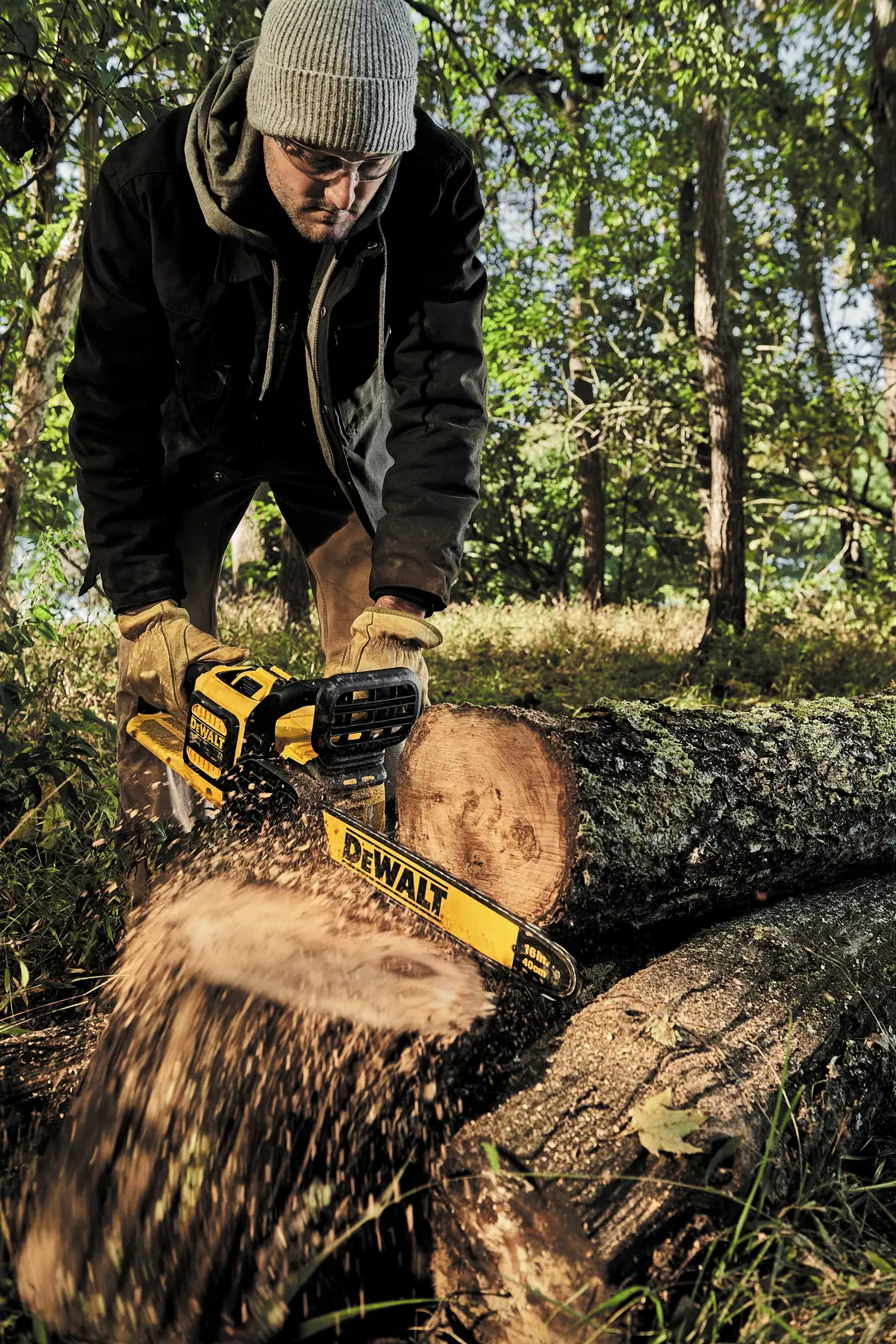 A person wearing gloves and outdoor clothing uses a DEWALT cordless chainsaw to cut a log in a forest setting. Sawdust flies as the chainsaw operates.