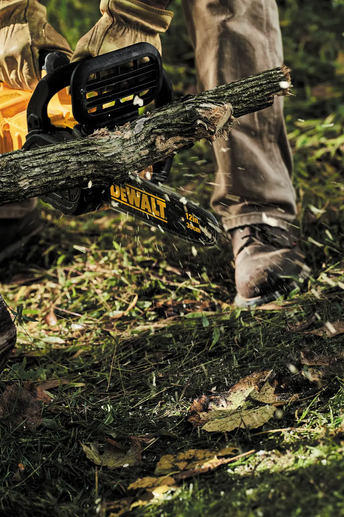 A person using a DEWALT 12-inch cordless chainsaw to cut a tree branch outdoors while wearing work gloves and boots.