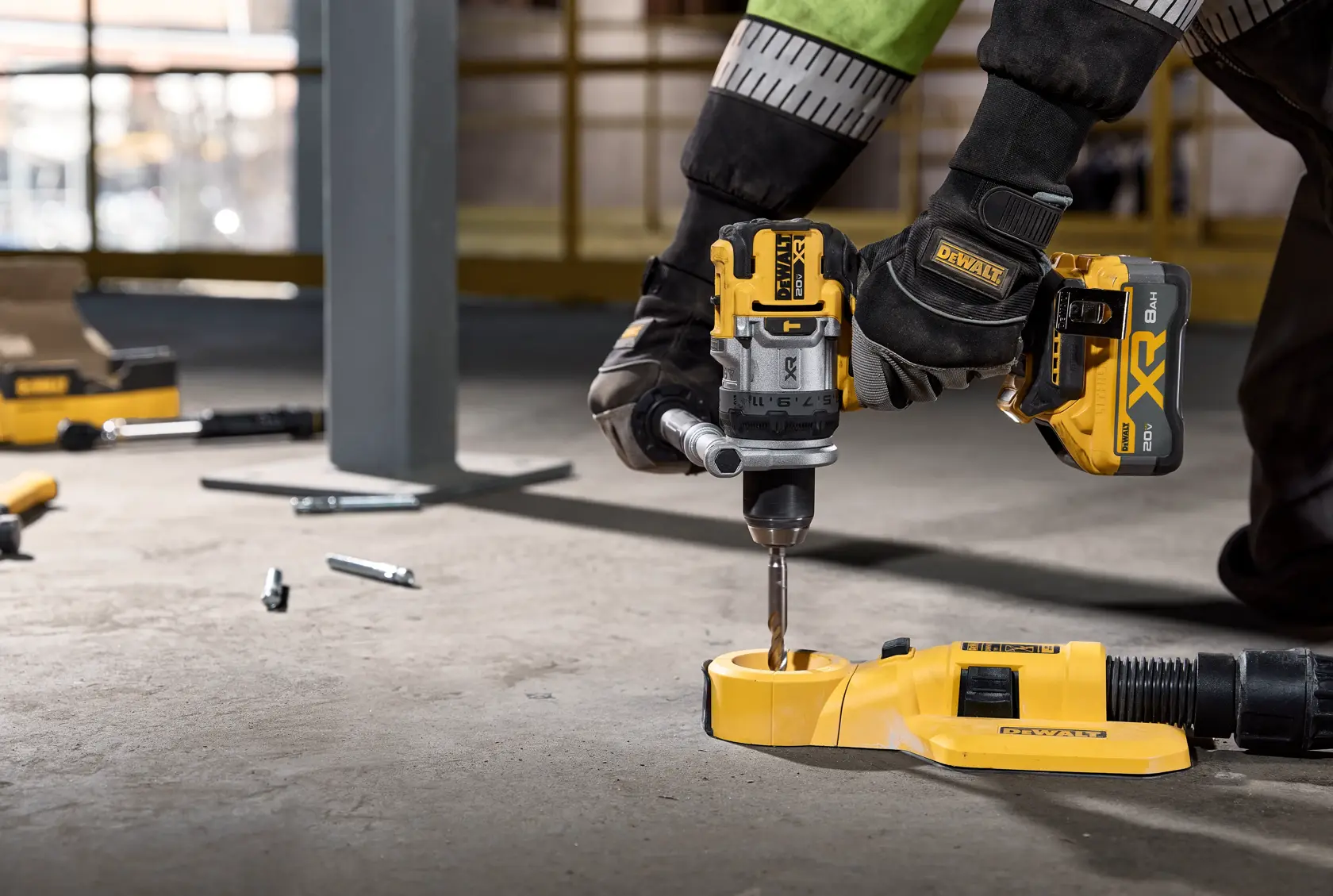A person wearing safety gloves is using a DEWALT cordless power drill to drill into a metal surface on the floor. Various bolts and tools are scattered around, and a yellow dust extraction attachment is connected to the drill.