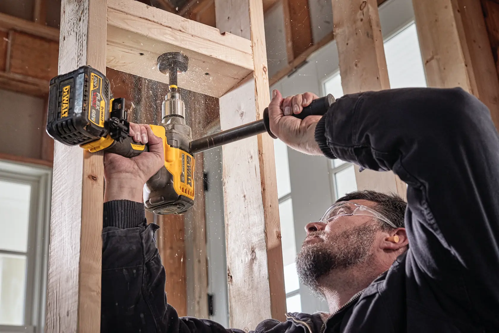 Person using a DEWALT cordless drill to bore a hole in wooden studs inside a building under construction. The drill is held with both hands and sawdust particles are visible.