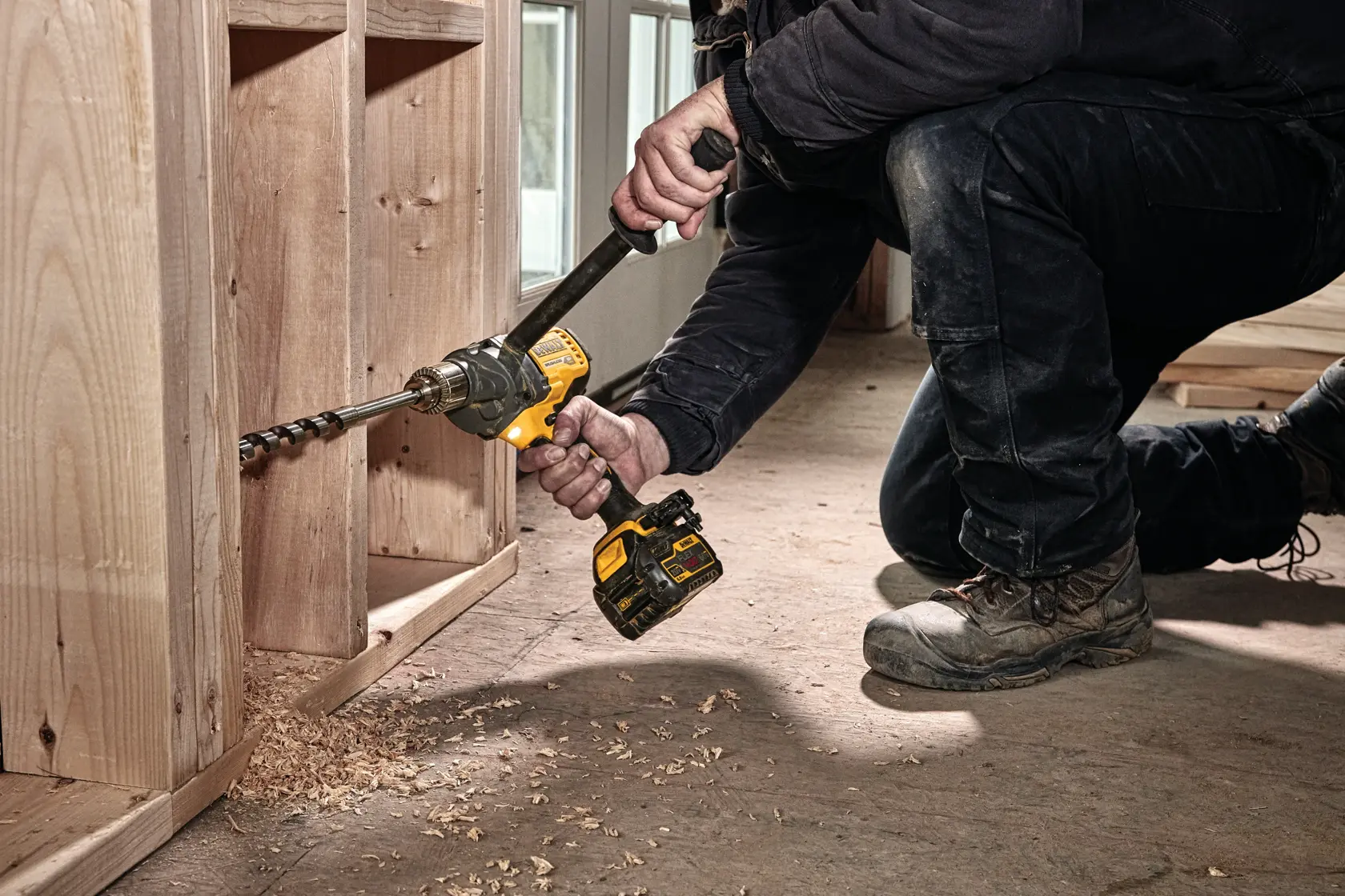 A person using a DEWALT cordless drill to bore a hole in a wooden structure, with wood shavings scattered on the floor.