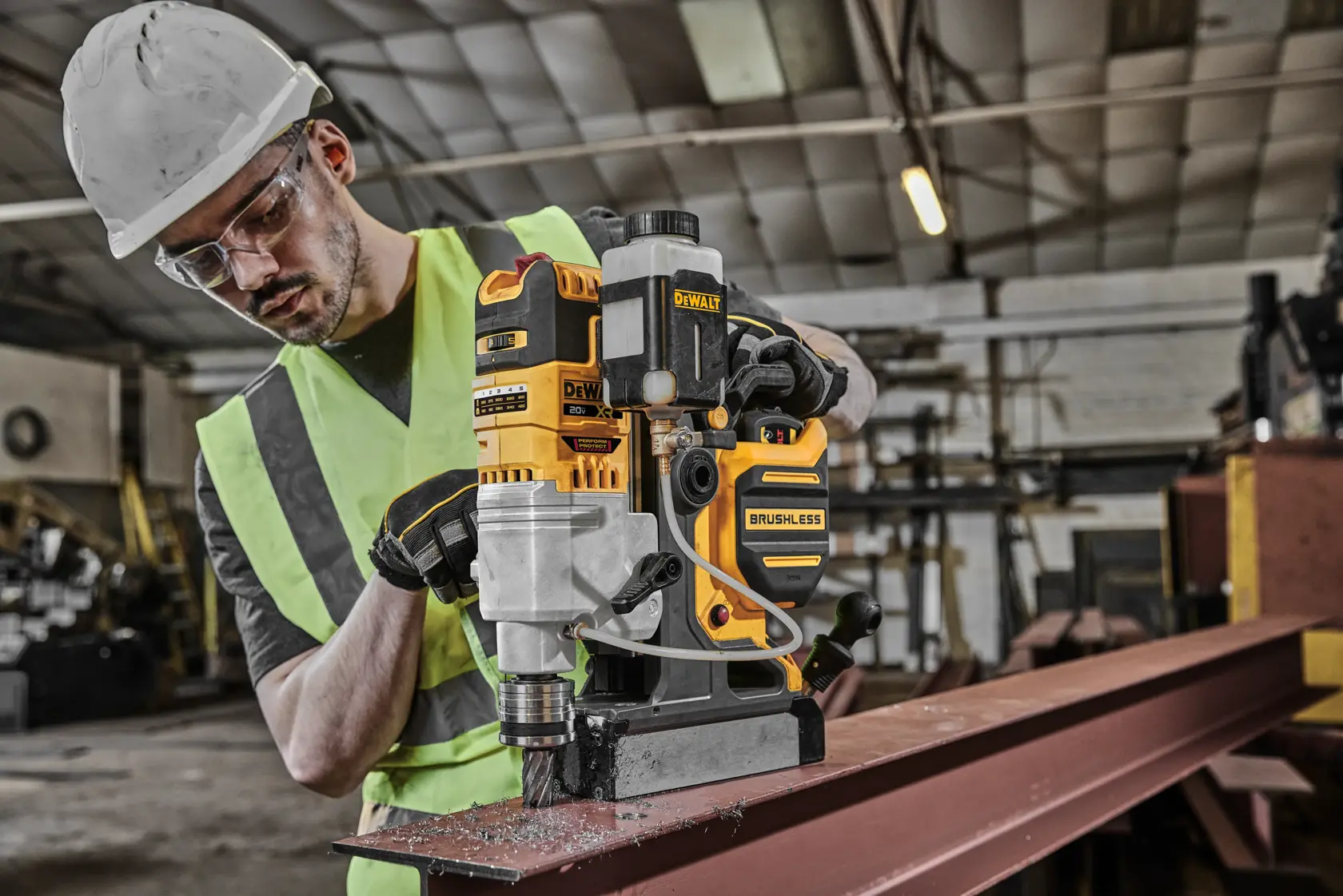 A worker wearing a safety helmet and reflective vest operates a DEWALT brushless magnetic drill (model DCD1624GX2) on a steel beam inside an industrial workshop.