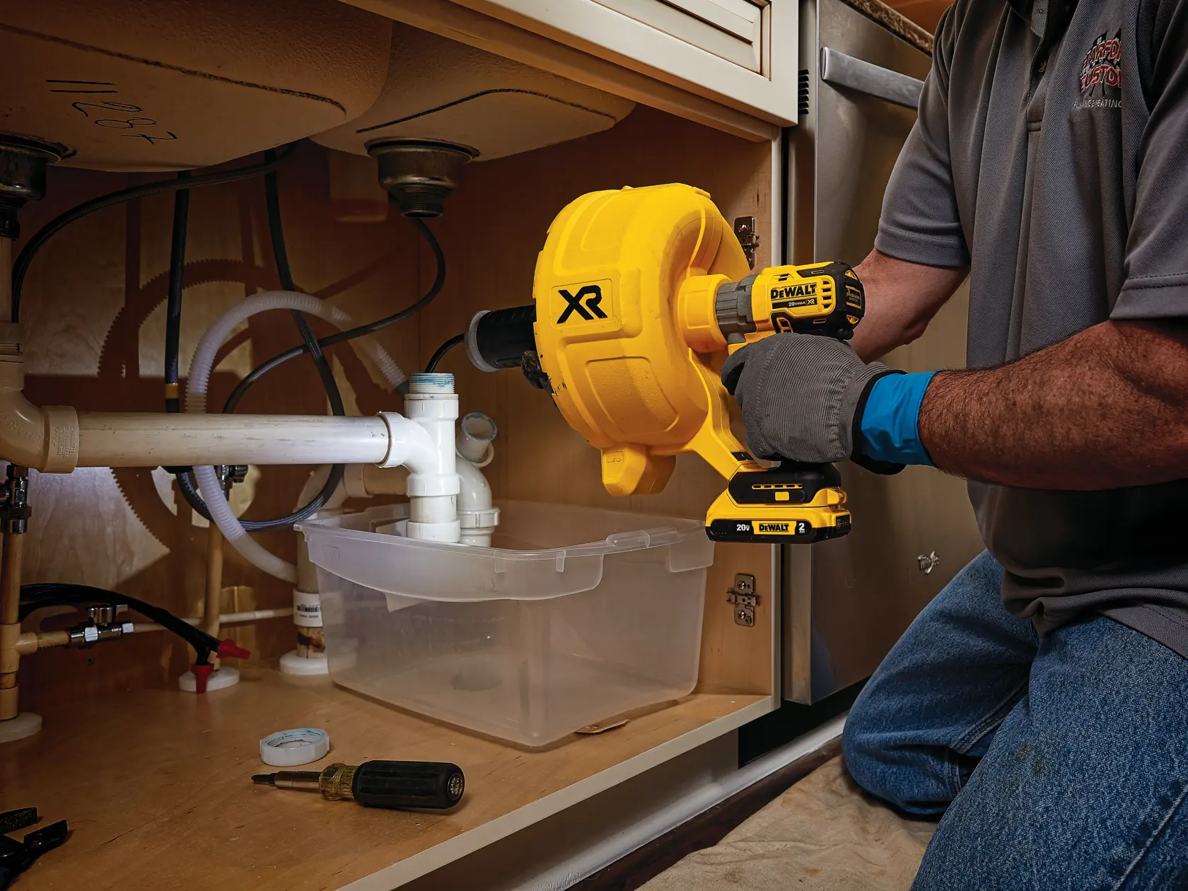 A person is kneeling and using a DEWALT DCD200D1 power tool to unclog pipes under a kitchen sink. The tool is yellow and black, and the person is wearing gloves and a gray shirt. There is a plastic container and screwdriver placed under the pipes.