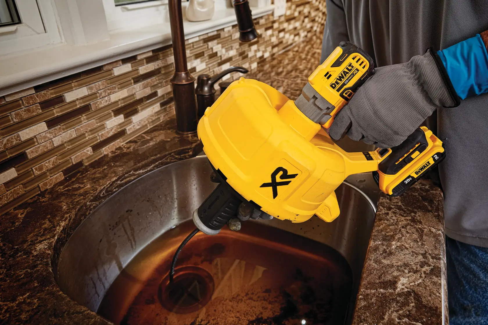 Person using a yellow DEWALT DCD200D1 cordless drain snake tool to unclog a pipe under a kitchen sink. The tool is operated inside a cabinet, with a plastic container placed under the pipes and various plumbing tools nearby.