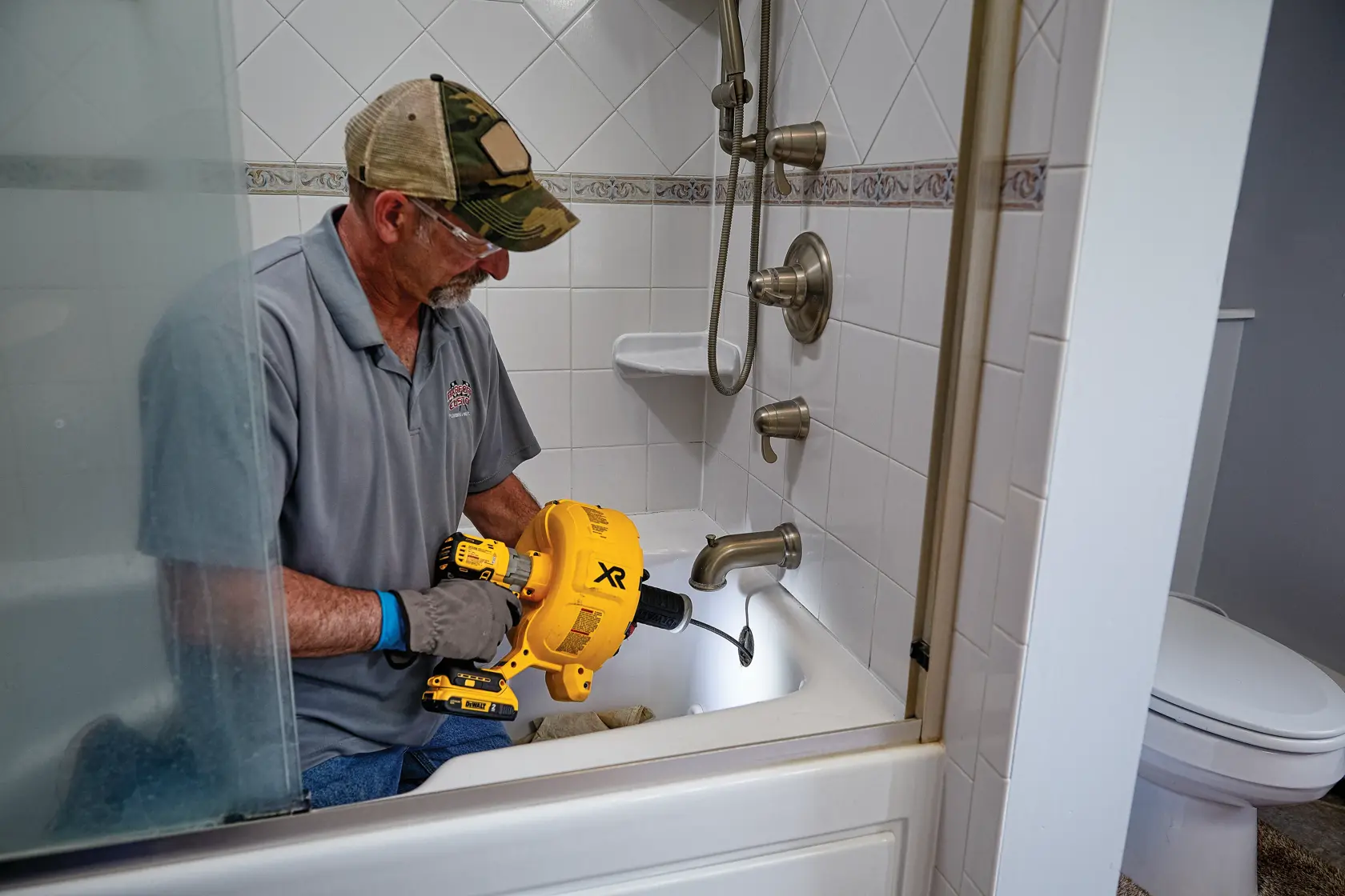 A person wearing gloves uses a DEWALT DCD200D1 power tool to unclog a kitchen sink drain. The yellow tool is inserted into the drain, and the sink is filled with dirty water.