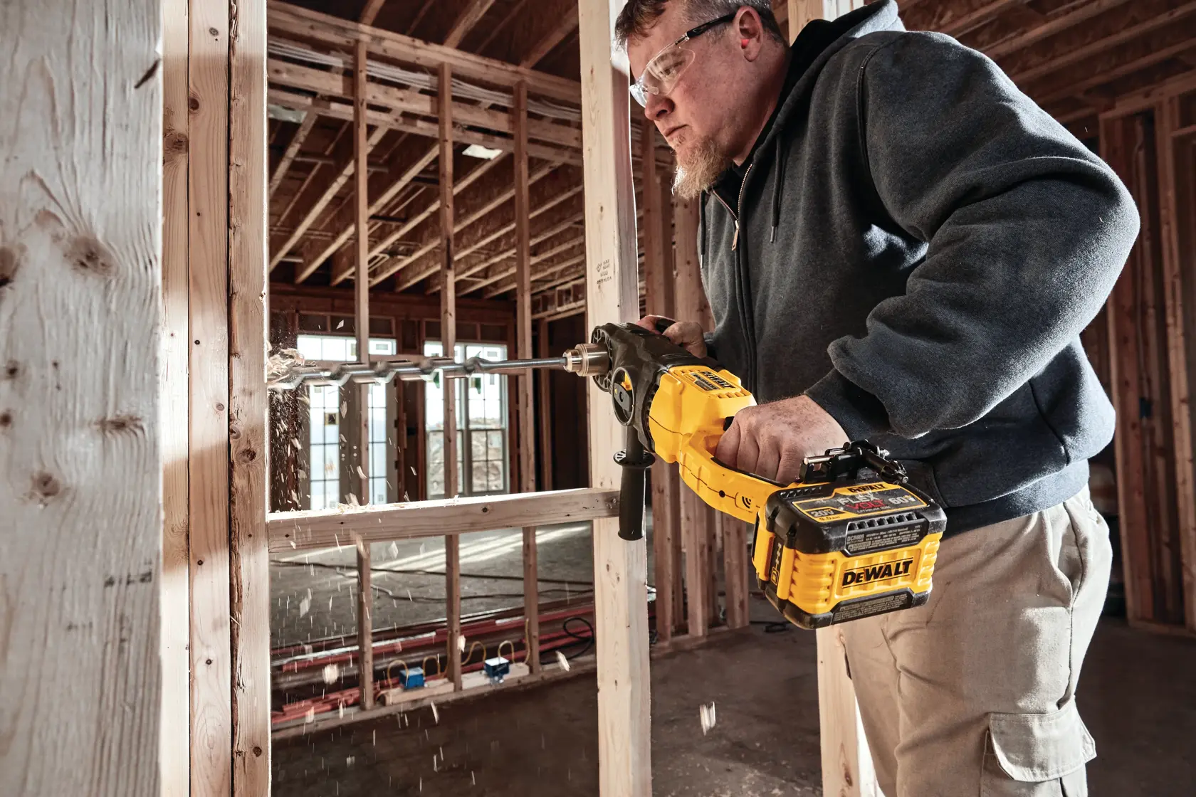 A person using a DEWALT cordless power drill to bore through wooden framing in a construction site. Wood shavings are visible as the drill operates.