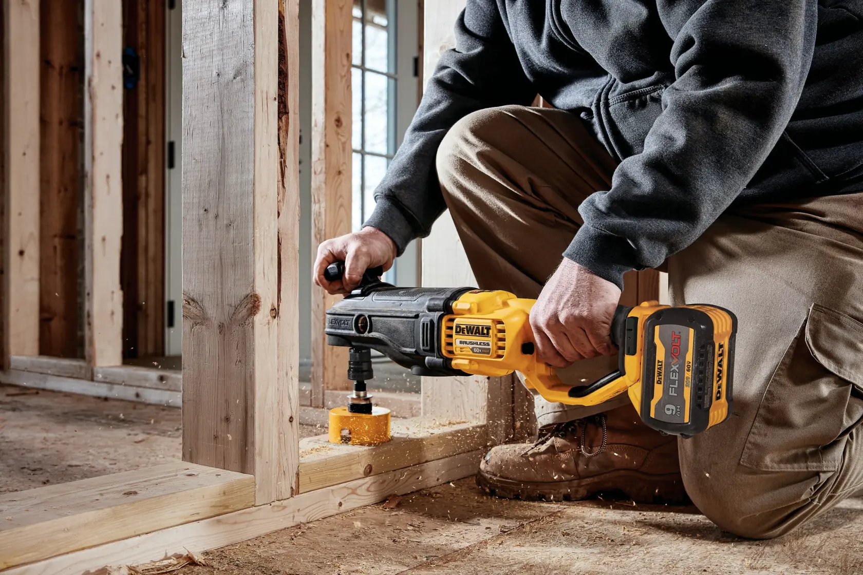 A person using a DEWALT cordless drill with a hole saw attachment to drill into a wooden frame inside a construction site.