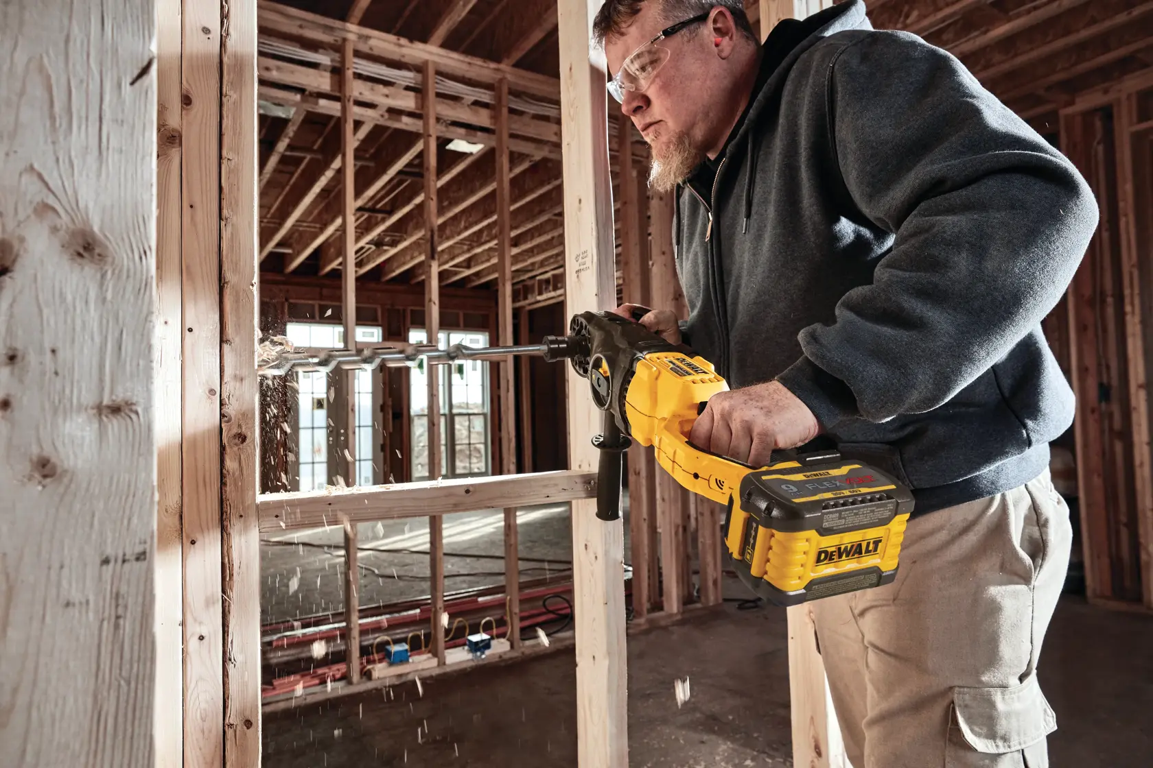 A person is using a DEWALT cordless drill to drill into a wooden frame inside a construction site. Sawdust is visible as the tool works, and the background shows unfinished wooden walls and beams.