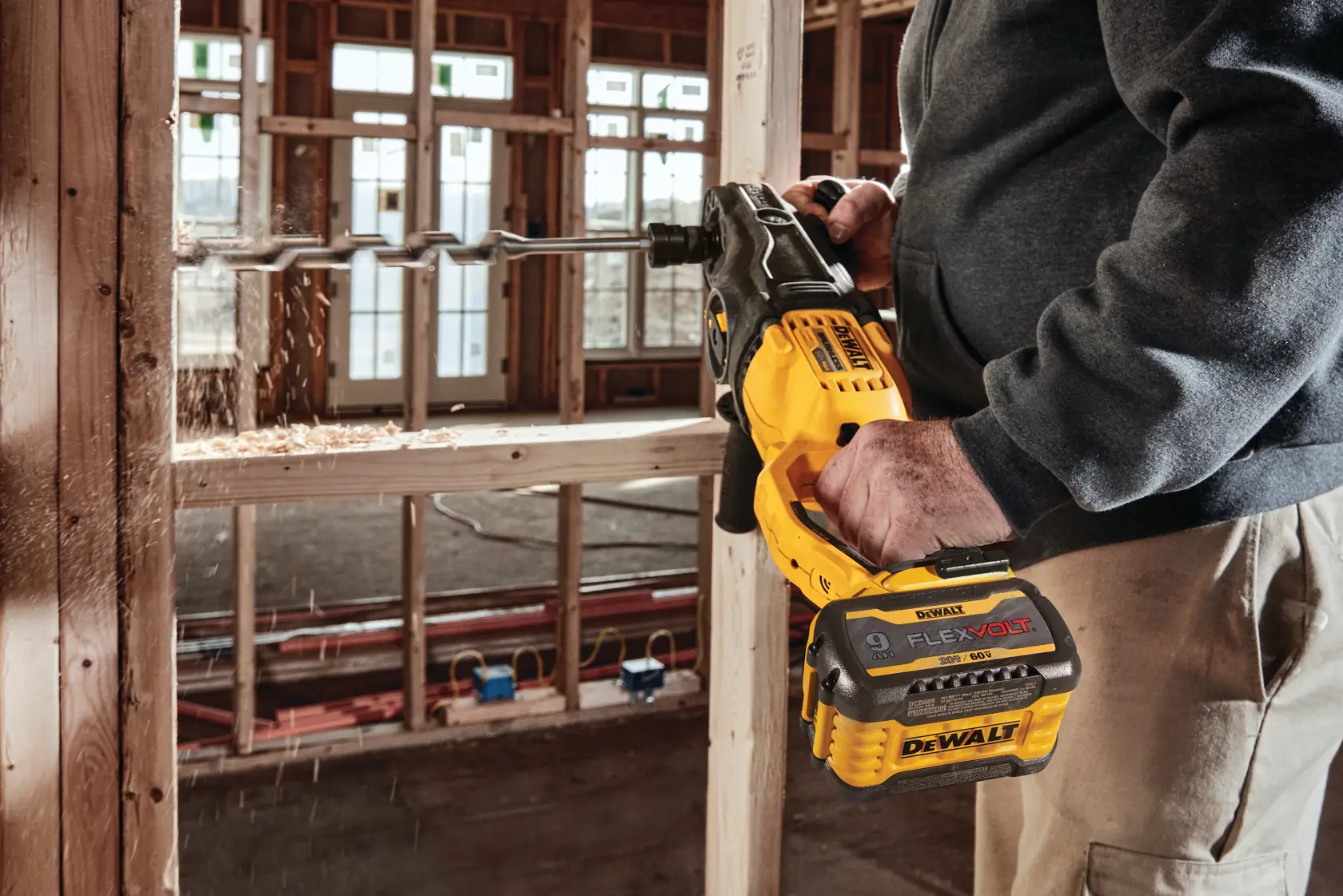 A person is using a DEWALT FlexVolt cordless drill with a large auger bit to drill into a wooden frame at a construction site.