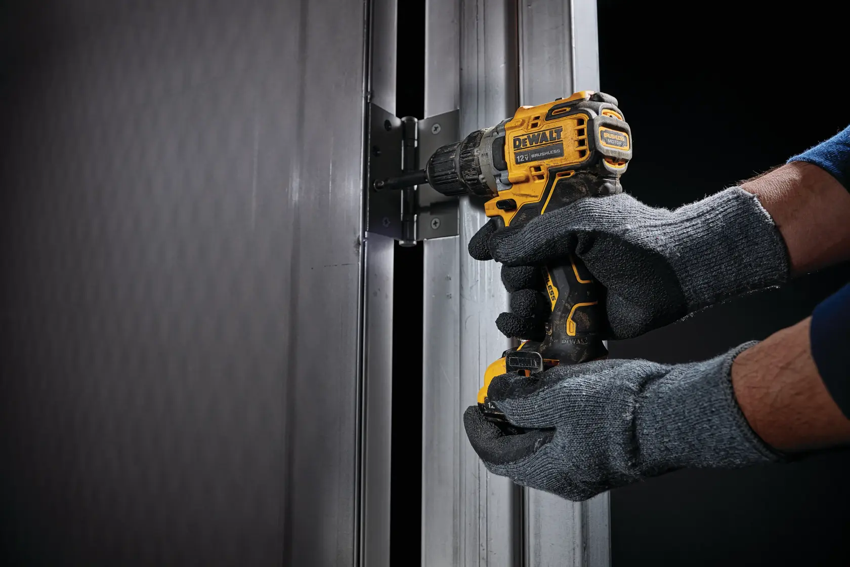 A person wearing grey gloves uses a DEWALT cordless drill to install a hinge on a metal door frame.