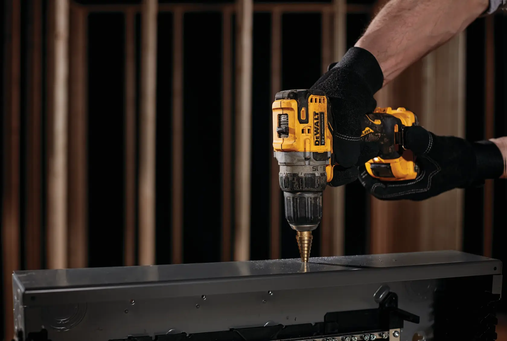 A person wearing black gloves uses a yellow DEWALT cordless drill to drill into a piece of metal.