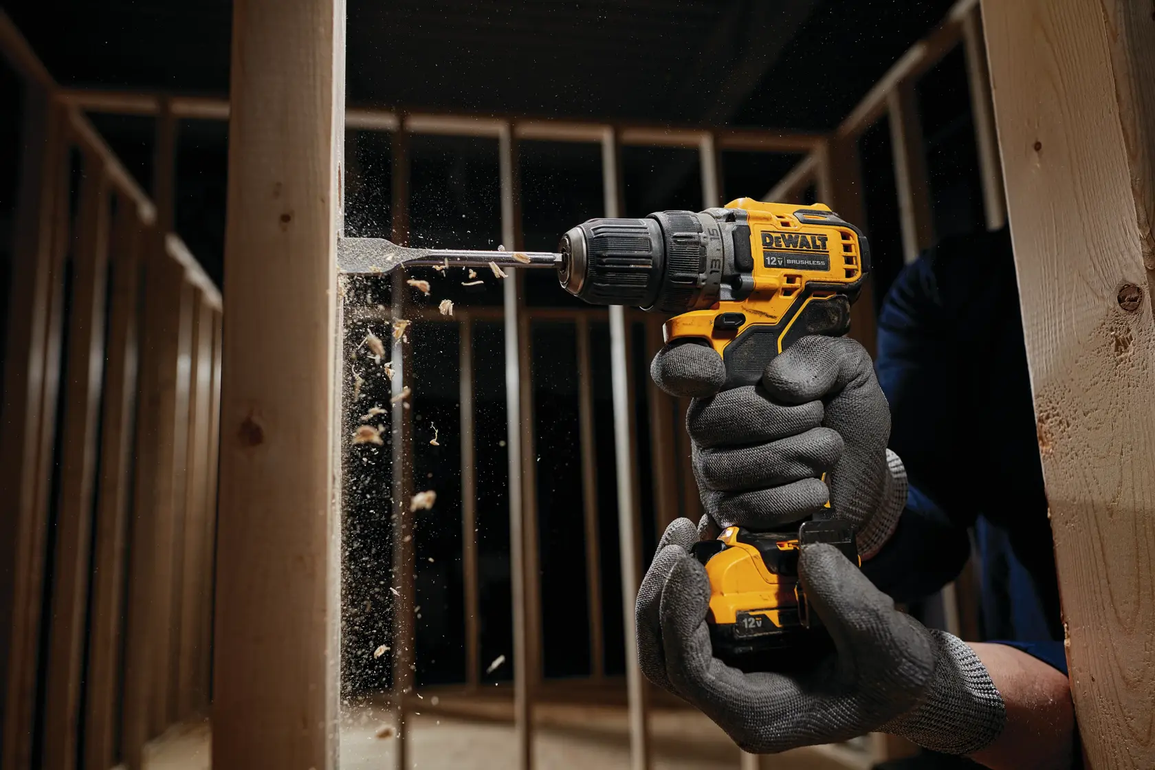 A person wearing protective gloves is using a DEWALT cordless drill to bore a hole in a wooden beam, with wood shavings flying, inside a construction frame.
