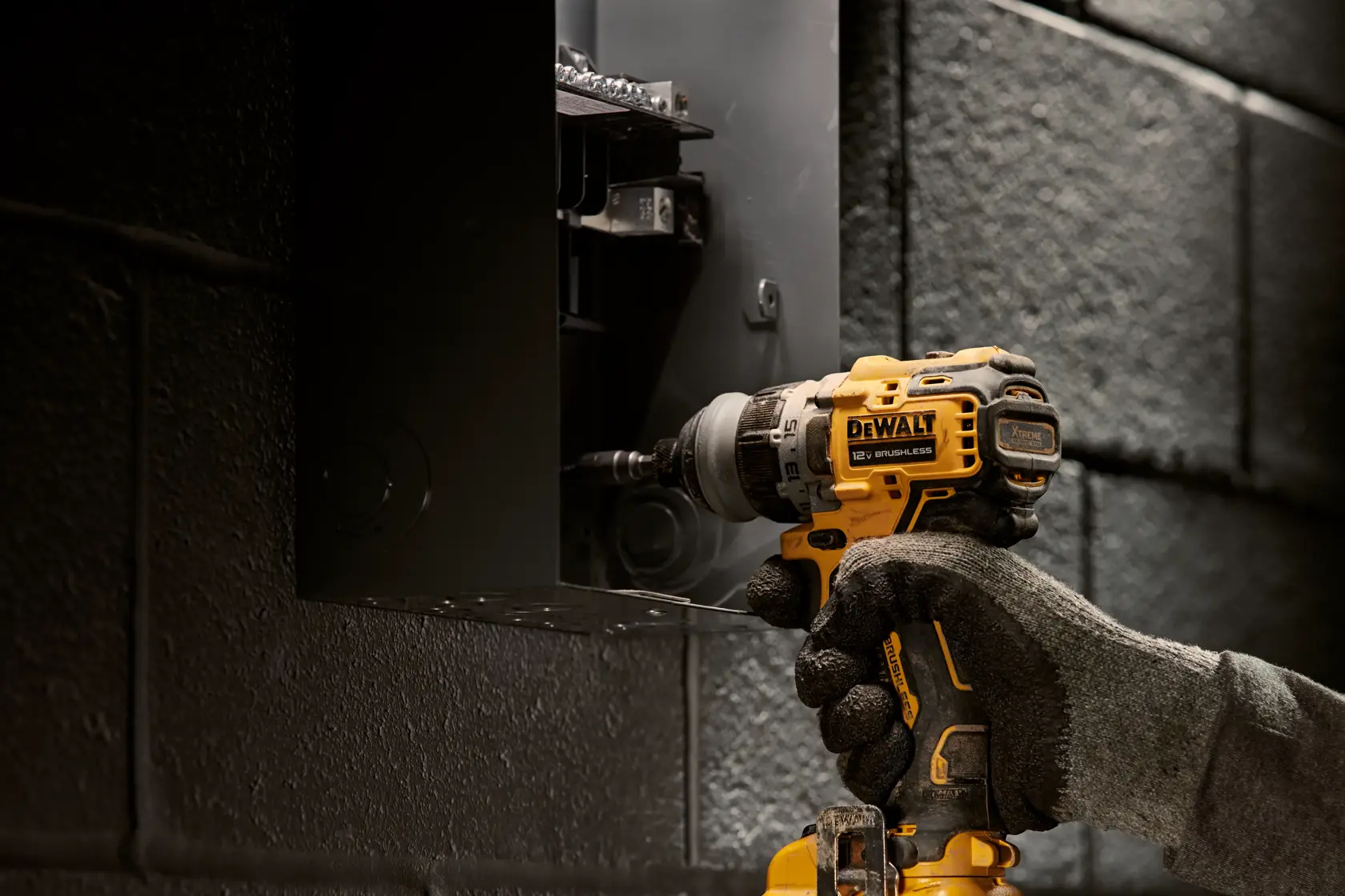 A gloved hand operates a DEWALT cordless drill, model DCD703F1, while working on a black electrical box mounted on a textured wall.