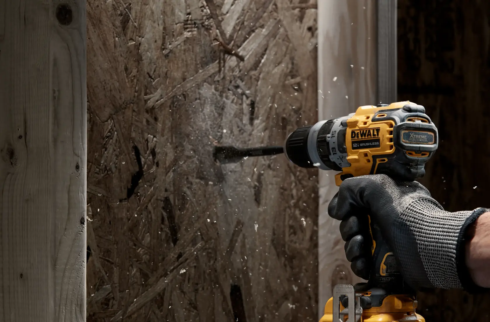 A person wearing protective gloves uses a yellow DEWALT cordless power drill to drill into a wooden board, with wood shavings flying from the drilling point.