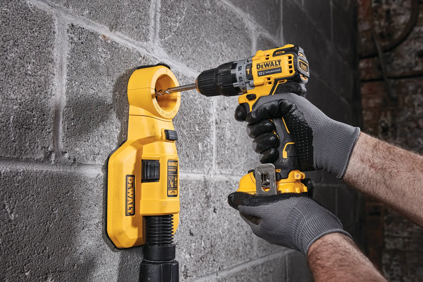 A person wearing gloves uses a DEWALT cordless drill to drill into a concrete wall, with a yellow DEWALT dust extraction attachment connected to the wall.
