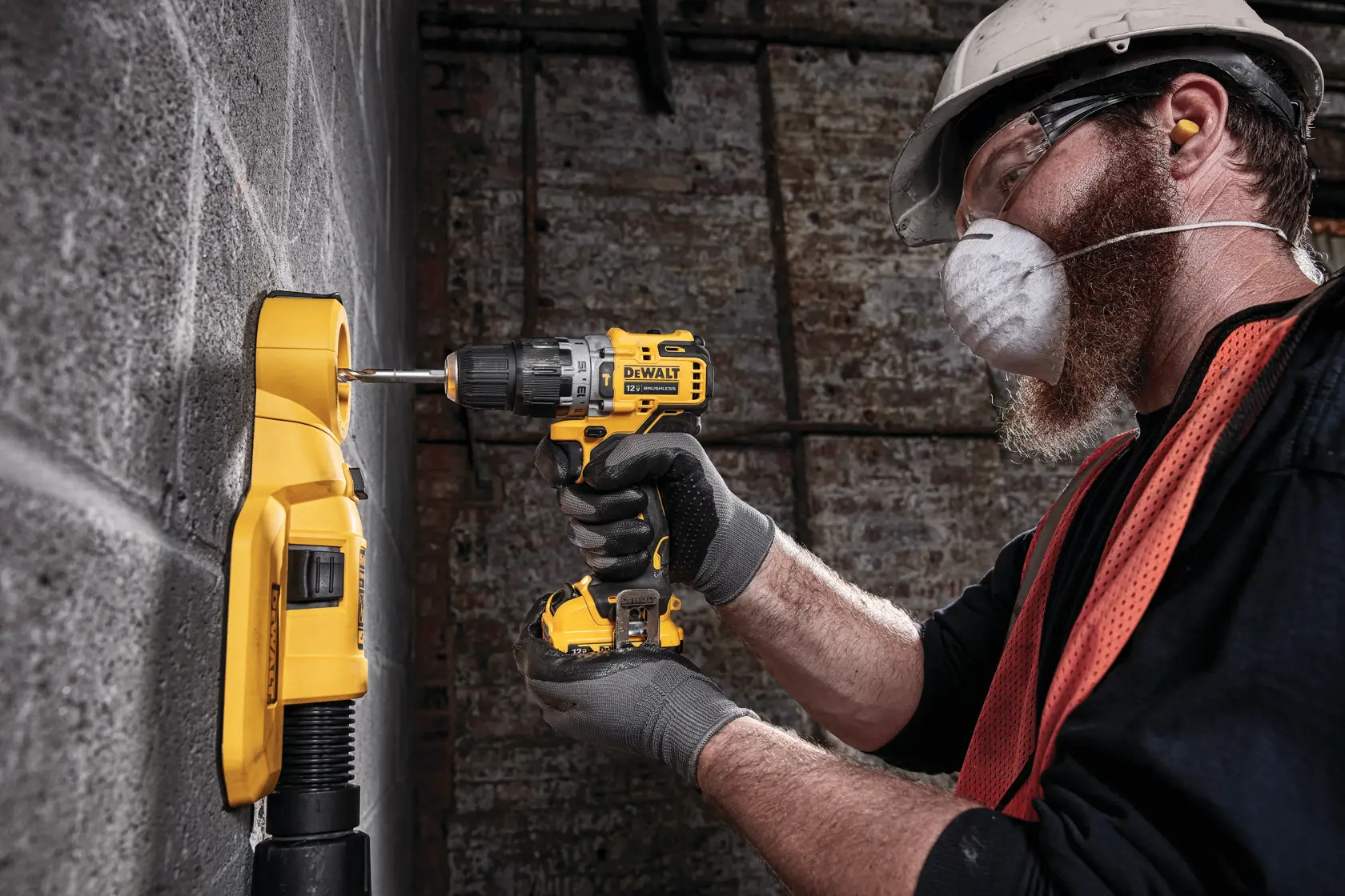 A person wearing safety gear uses a yellow DEWALT cordless drill to drill into a concrete wall, with a yellow dust extractor attached to the wall for collecting debris.