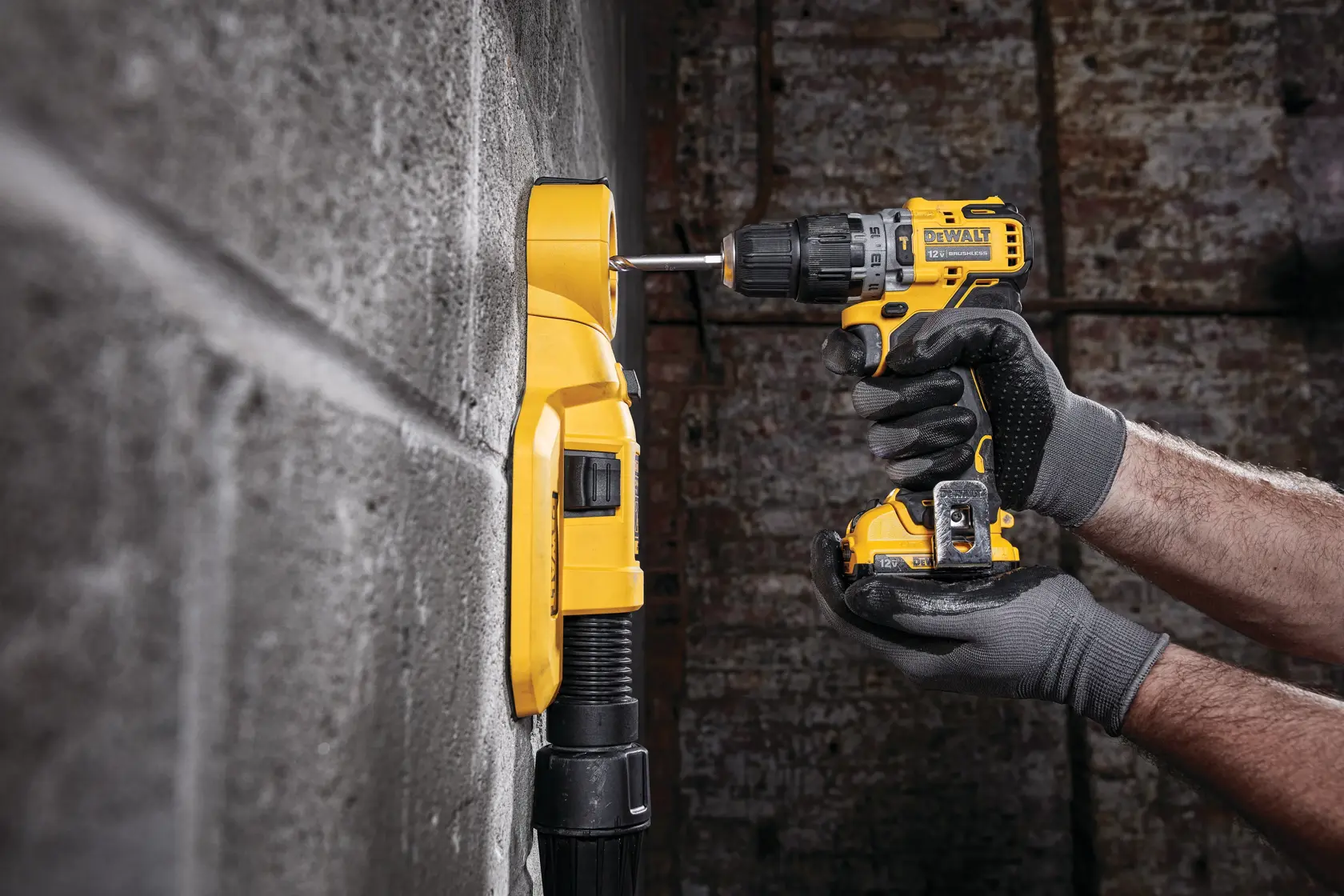 A person wearing gray work gloves is using a yellow DEWALT cordless drill to drill into a concrete wall. A yellow dust extraction device is attached to the wall to collect debris as the drilling takes place.