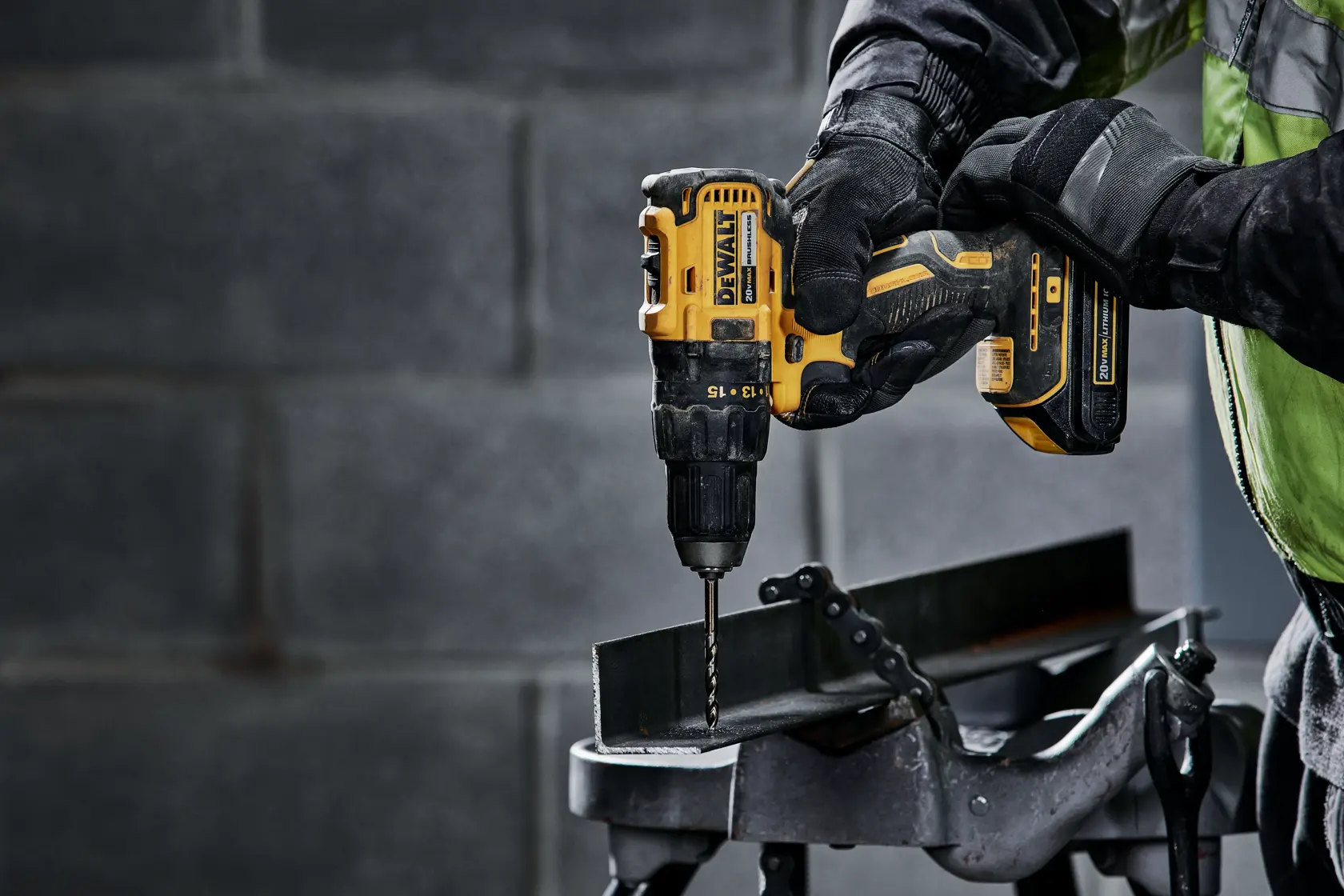 A close-up of a person using a DEWALT DCD777C2 cordless drill to drill into a metal beam in an industrial setting. The person is wearing gloves and a work jacket.