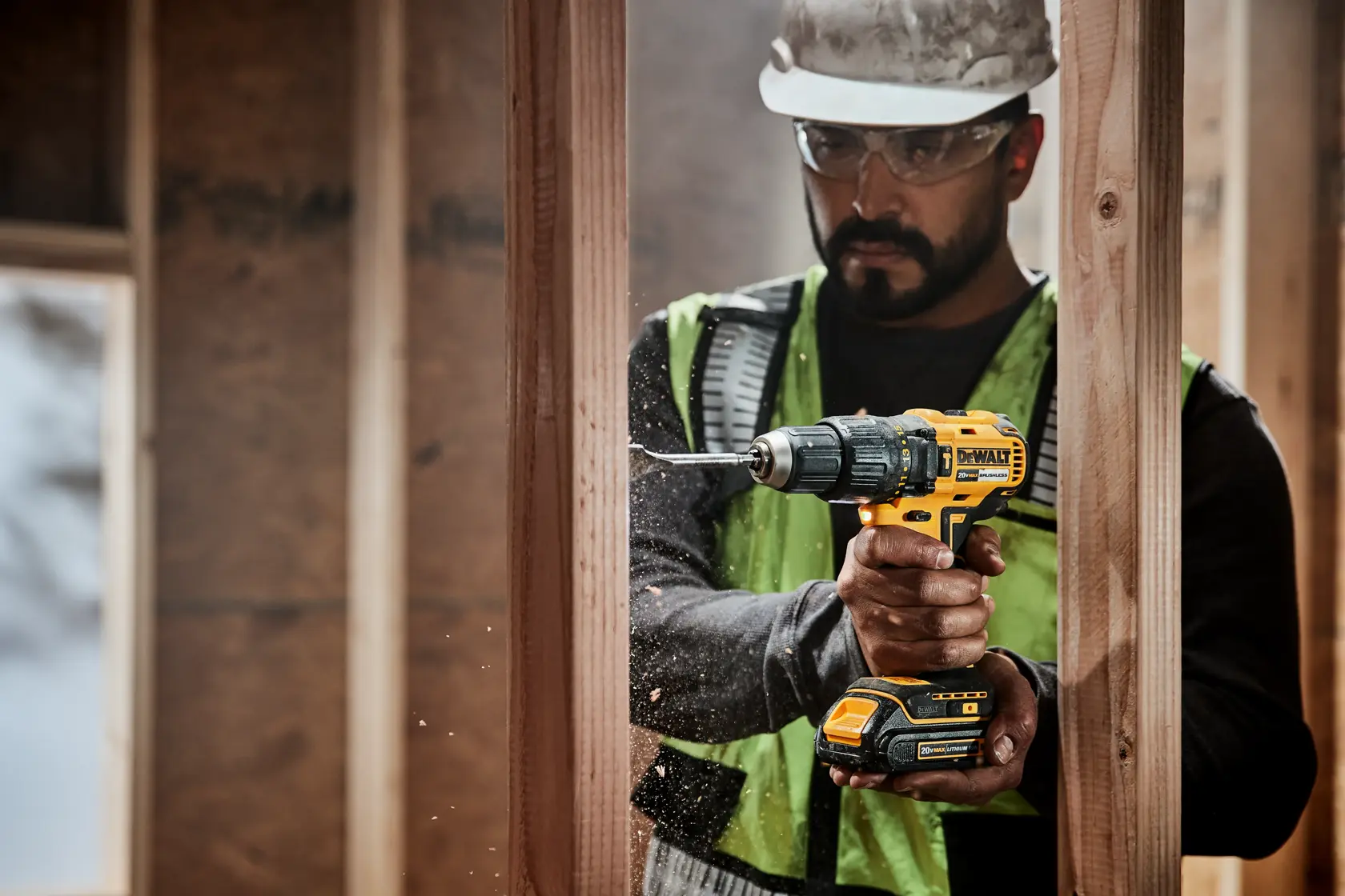 A person wearing a safety vest and helmet is using a DEWALT cordless drill to drill into a wooden frame at a construction site. The model DCD778C2 is visible on the drill. The person's face is obscured.