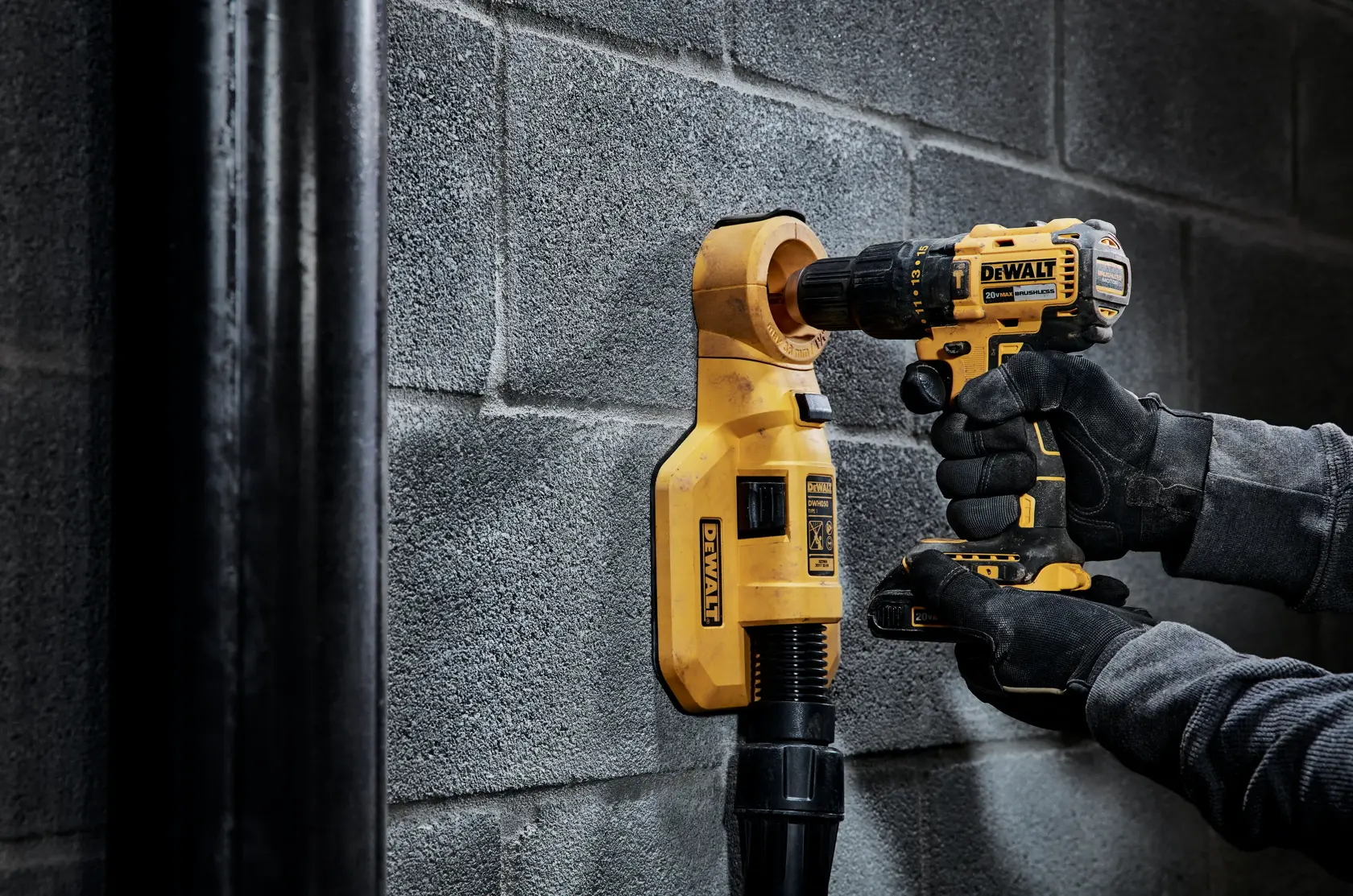 A person wearing gloves operates a DEWALT cordless drill to drill into a concrete wall, using a dust extraction attachment to collect debris.