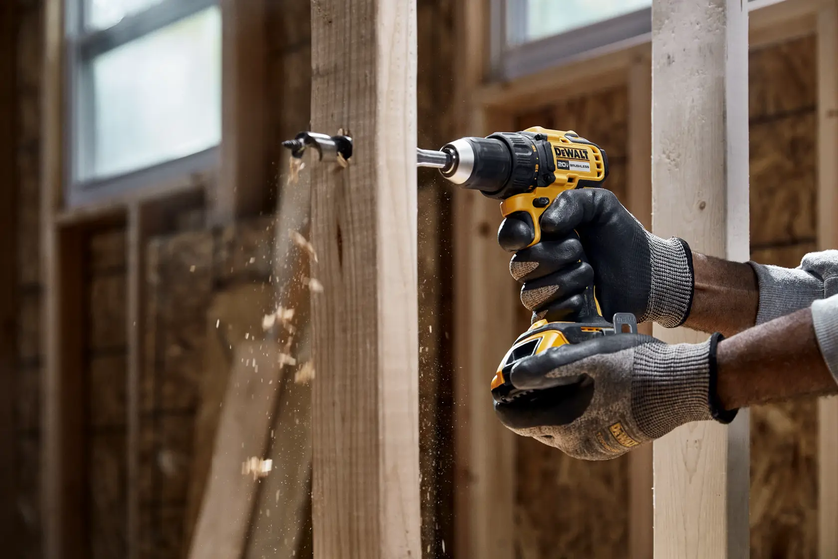 A person wearing gloves uses a DEWALT cordless drill to drill a hole into a wooden beam, with wood shavings flying in a construction environment.