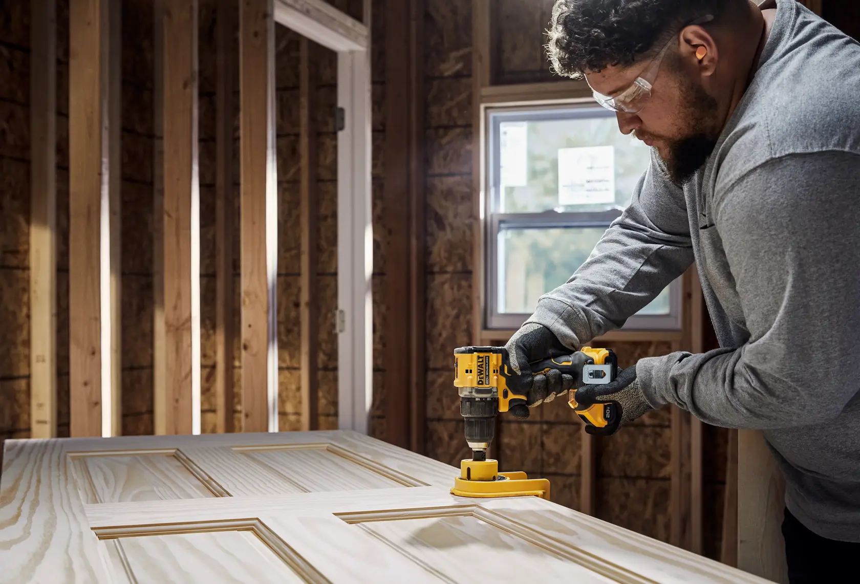 A person using a yellow DEWALT cordless drill to work on a wooden door in a construction setting. The person's face is blurred for privacy.