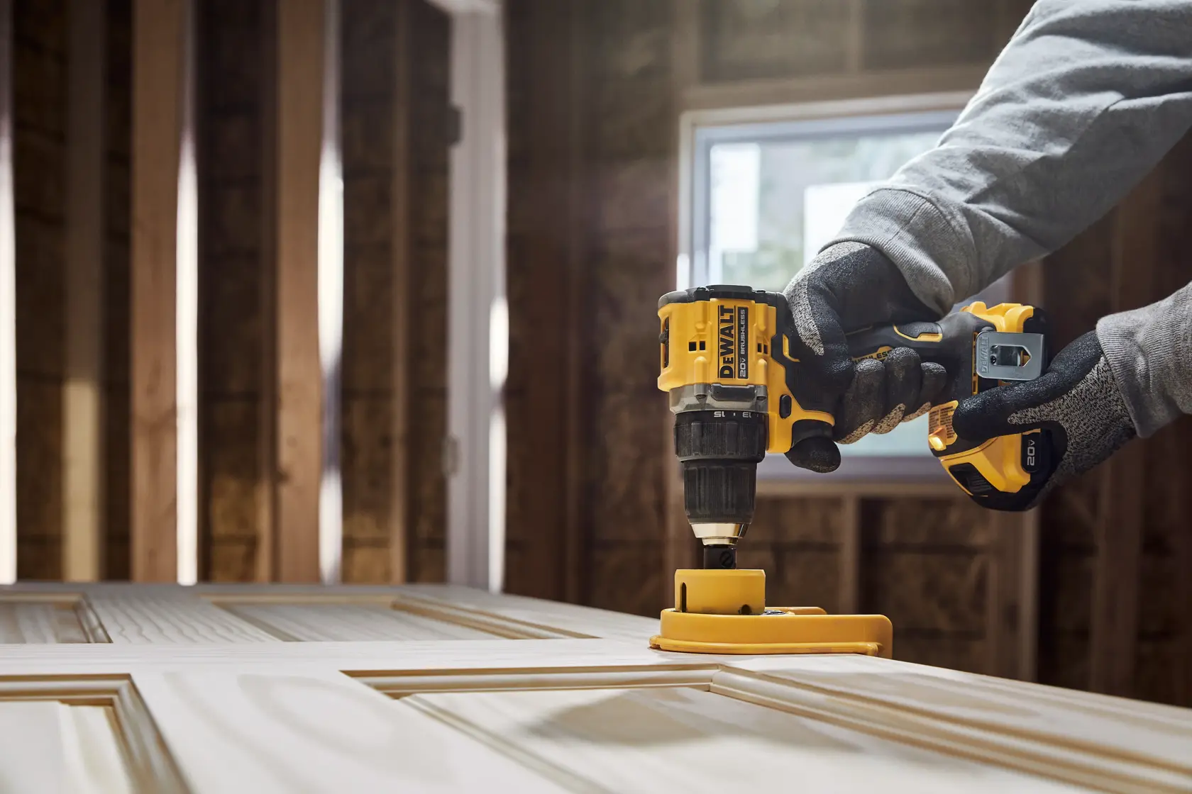 A person wearing gloves is using a yellow DEWALT cordless drill on a wooden door in a room under construction.